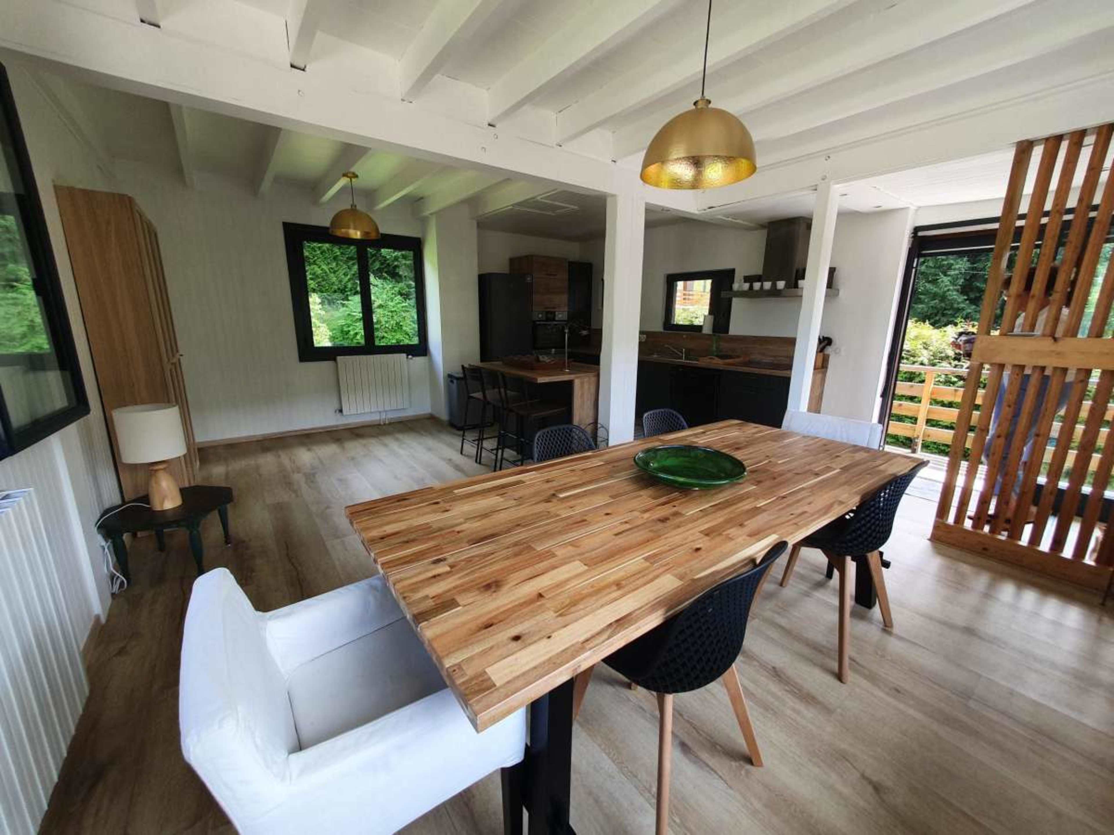 The image shows a modern kitchen and dining area featuring a wooden table surrounded by black chairs, with large windows providing natural light and access to a deck.