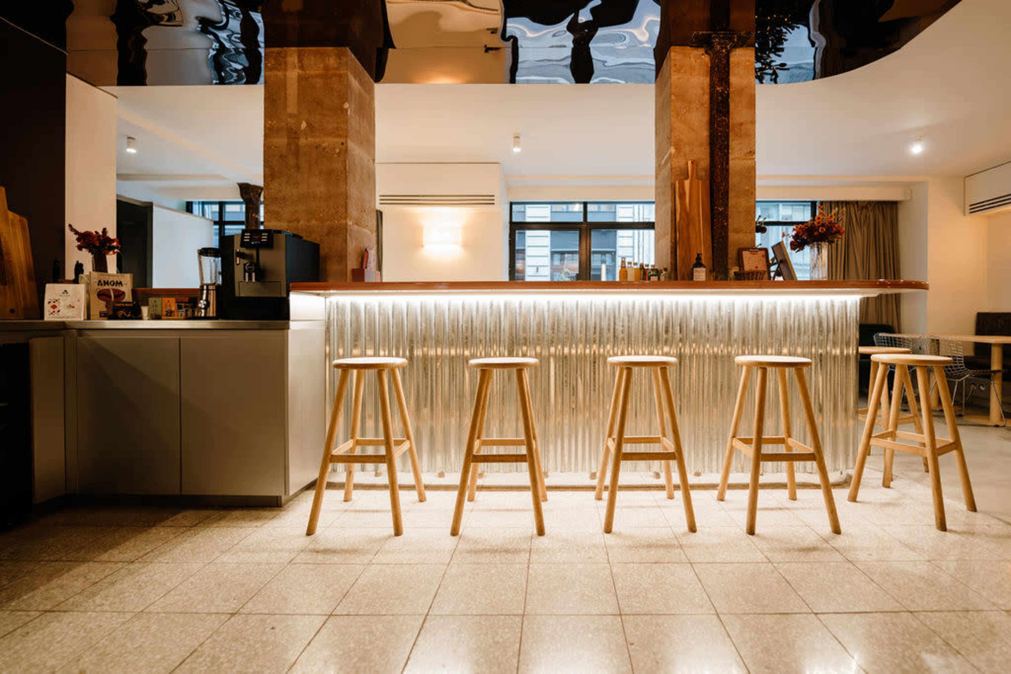 The image shows a modern bar area with a polished counter, wooden stools, and a stylish interior design featuring a reflective ceiling.