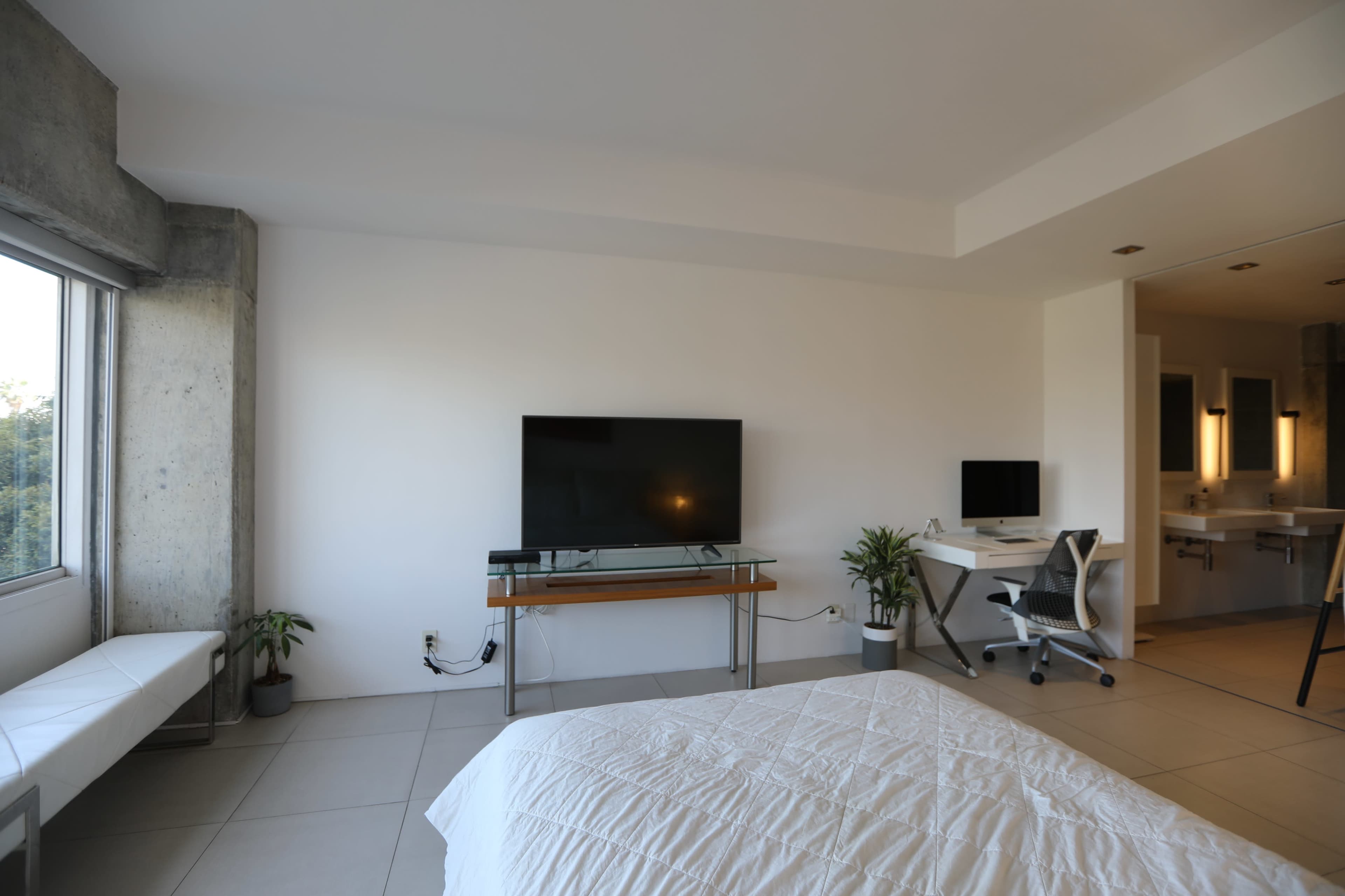 A modern bedroom features a bed, a mounted television on the wall, and a desk with a chair, adjacent to a large window.