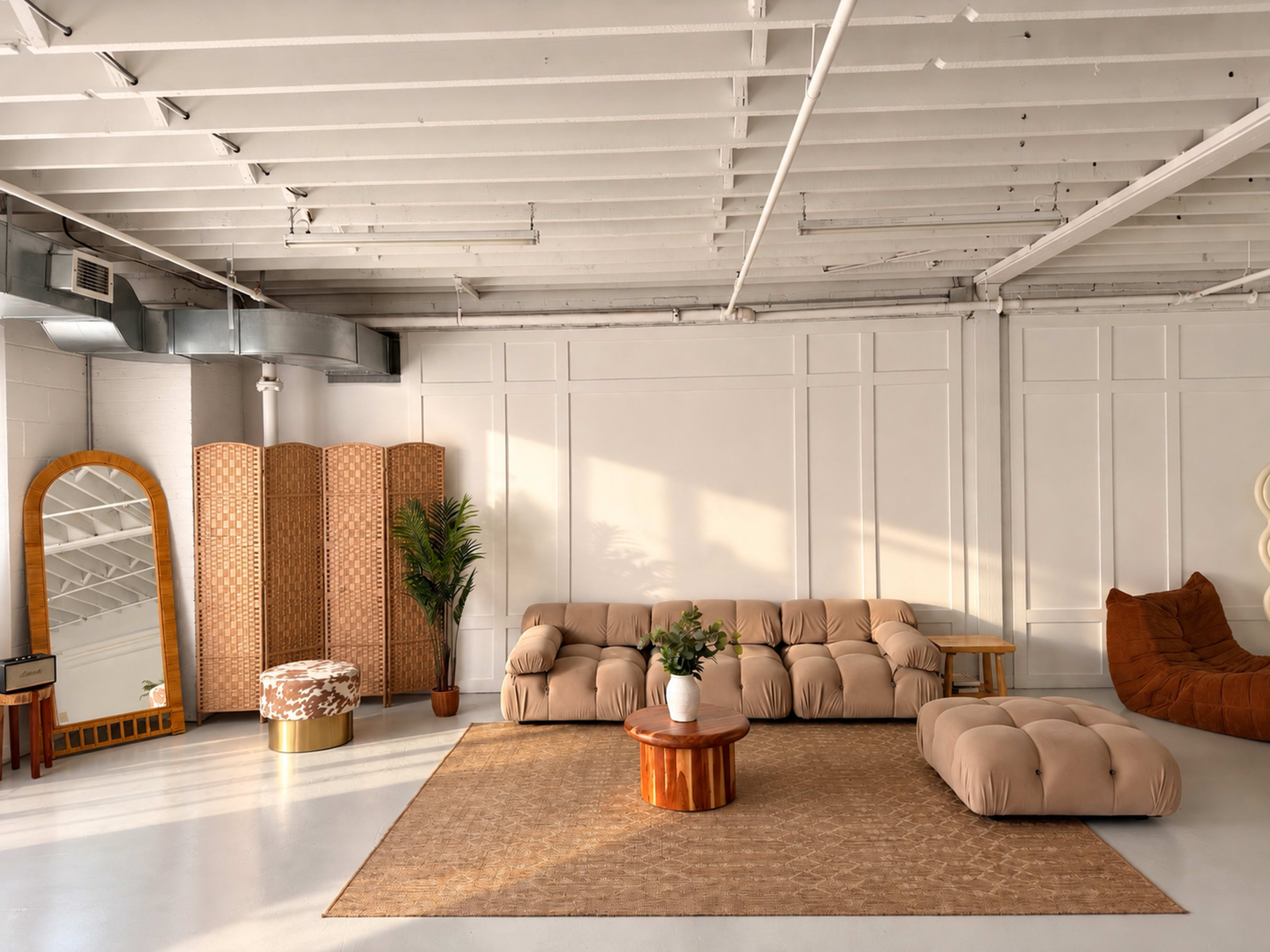The image features a spacious, well-lit living area with a beige sectional sofa, a circular wooden coffee table, decorative plants, and a large mirror against a white wall.