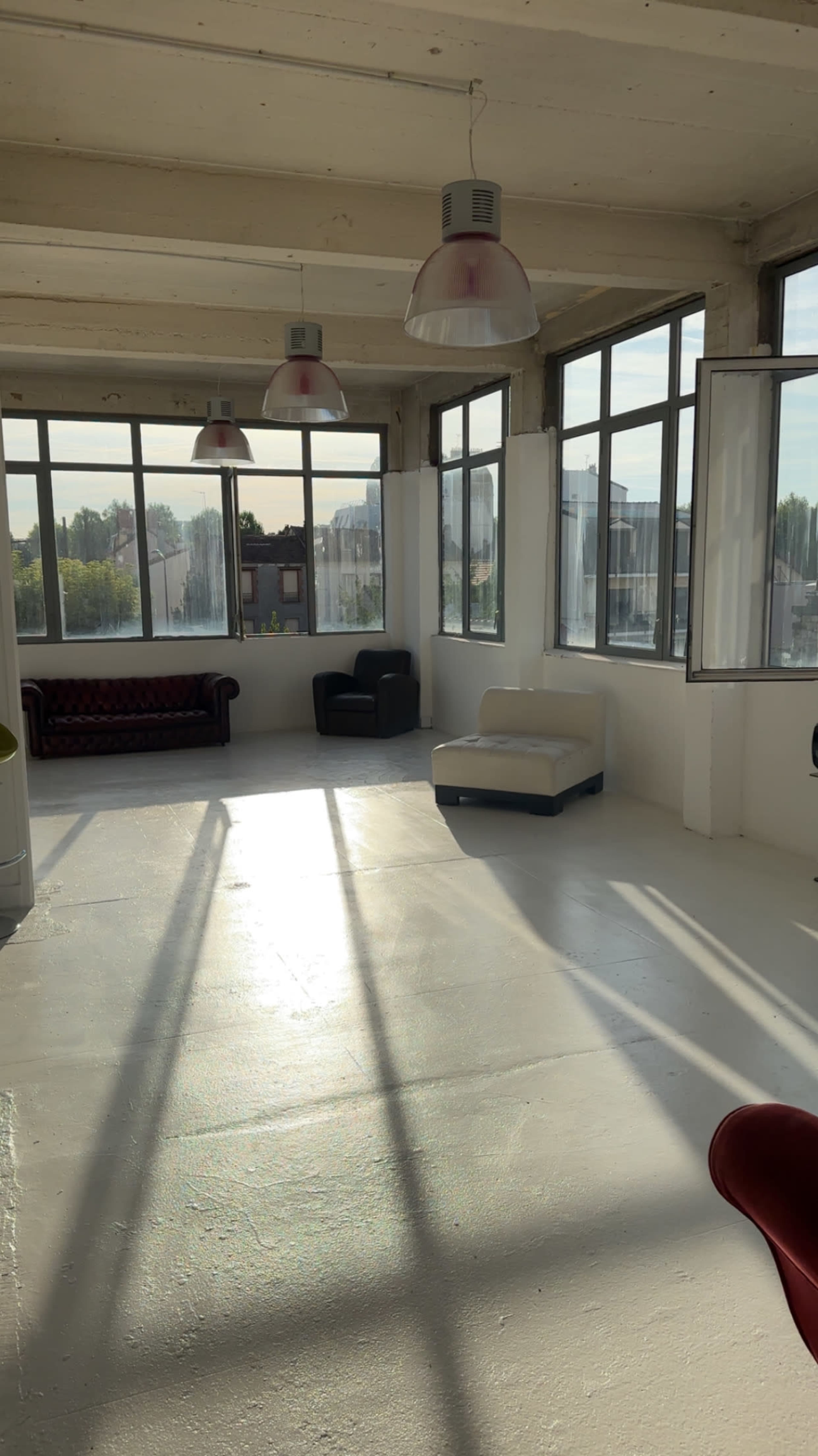 The image shows a spacious, well-lit room with large windows, featuring a few sofas and chairs arranged on a bare floor.