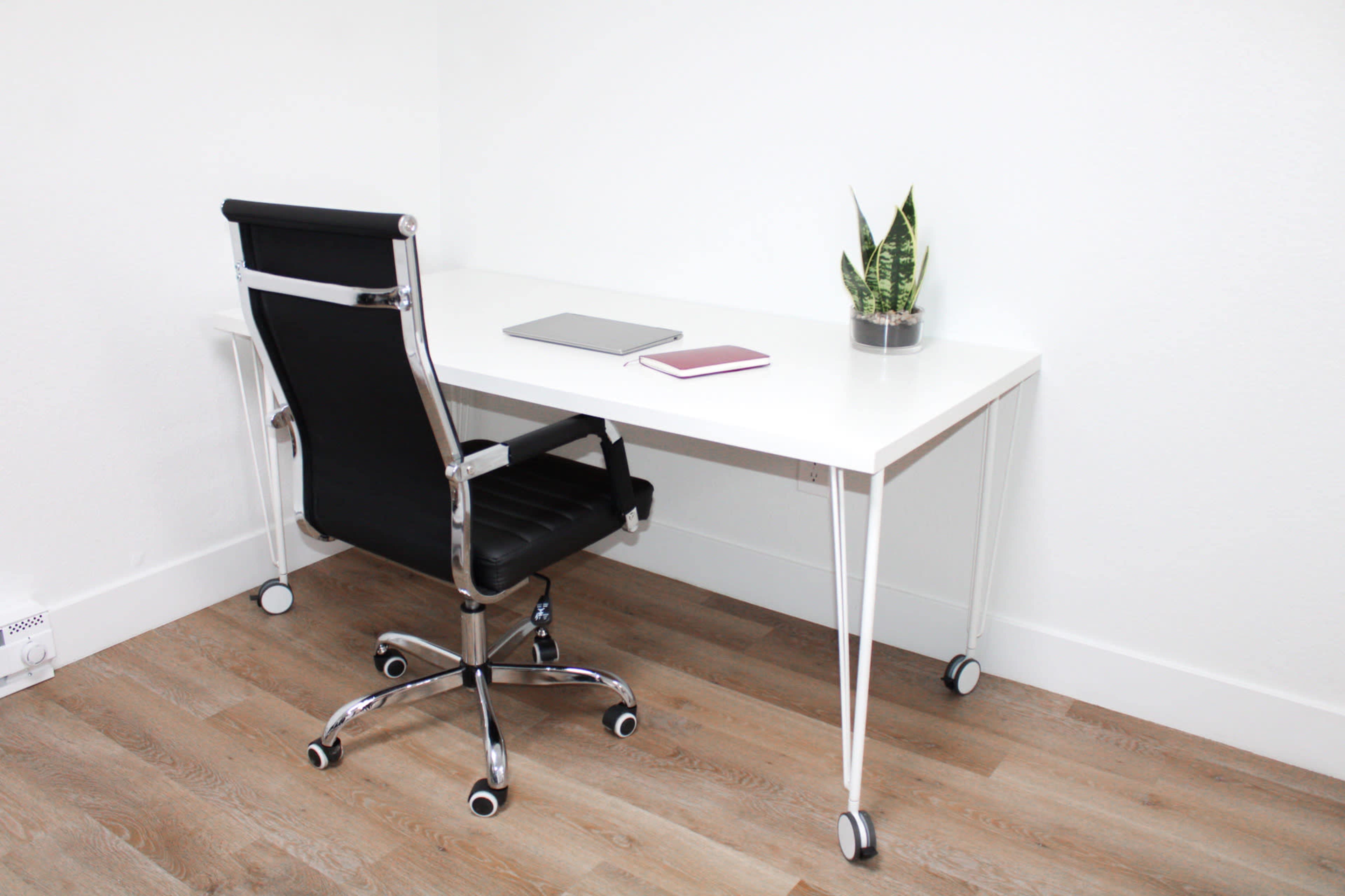 A minimalist office setup features a white desk, a black rolling chair, a laptop, a notebook, and a potted plant.