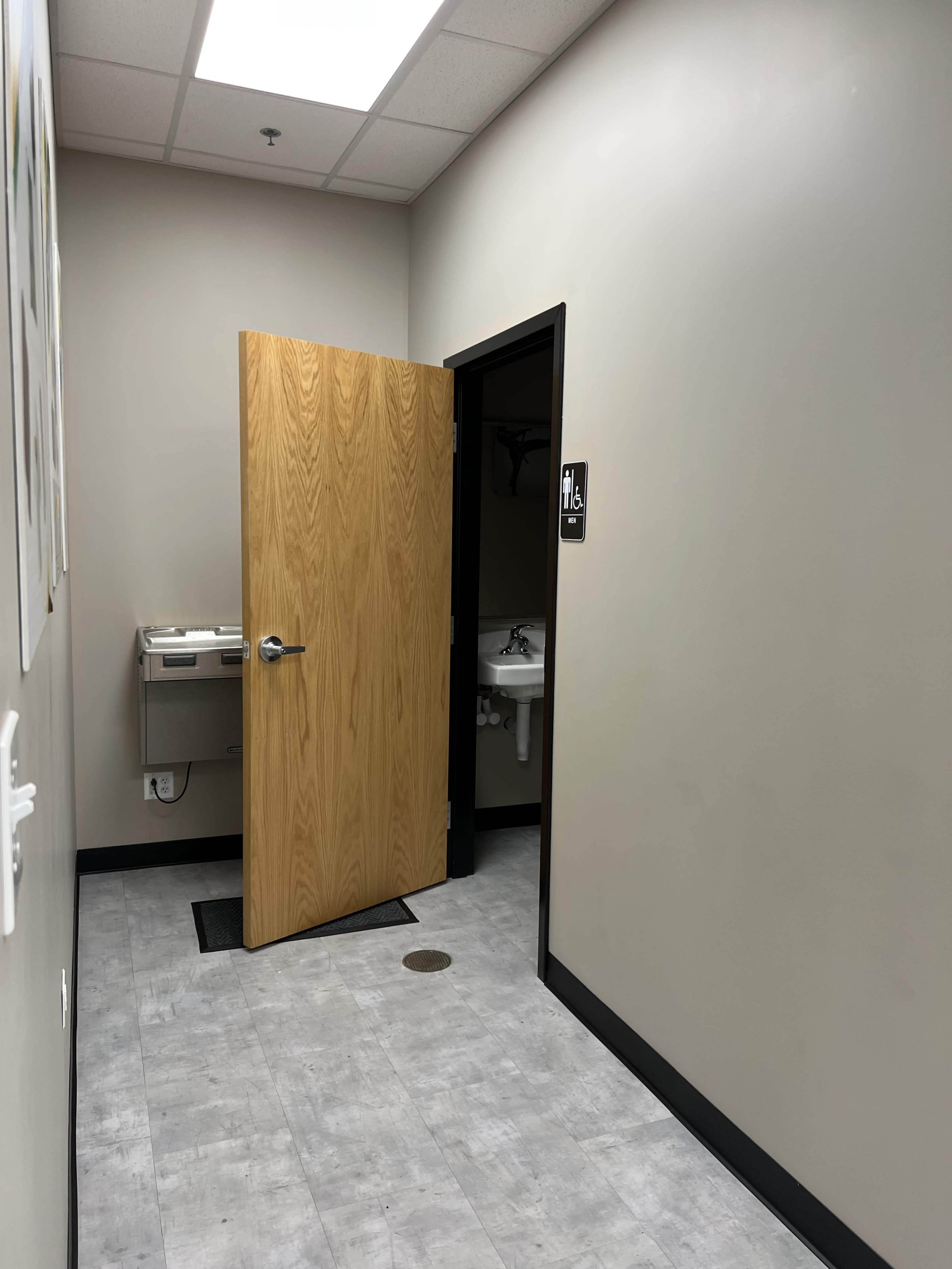 The image shows a hallway with a wooden door partially open on the right, leading to a restroom, and a handwashing sink visible.