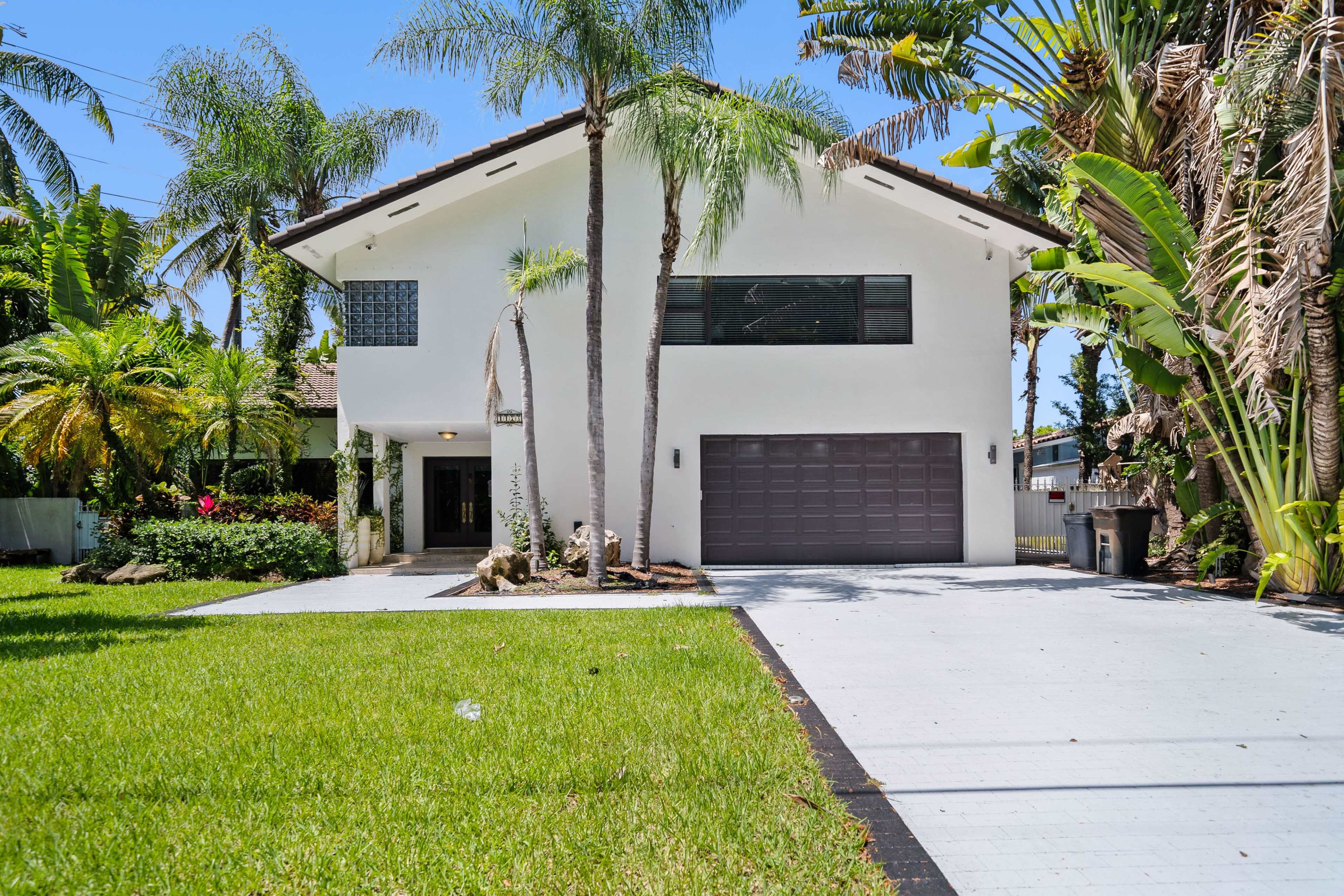 A modern two-story white house with a gray garage door, surrounded by palm trees and lush greenery.