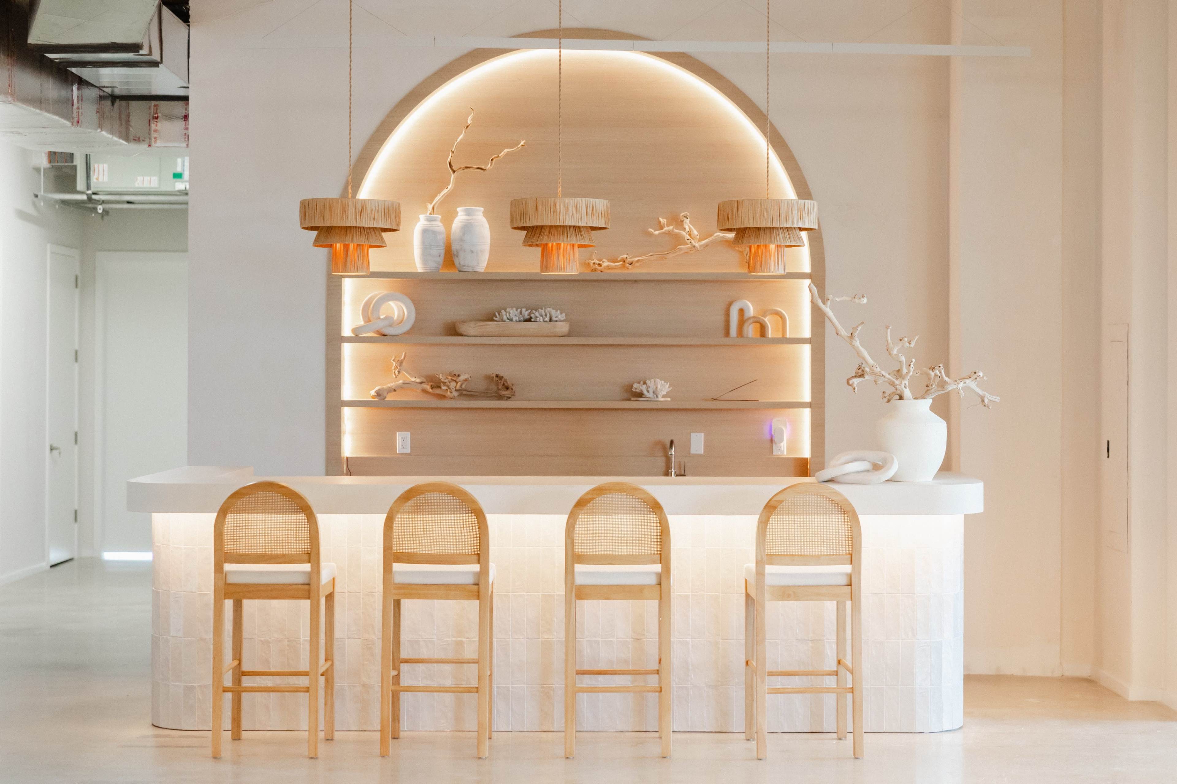 A modern minimalist bar features a curved wooden backdrop with shelves adorned with decorative items and three unique pendant lights above a white countertop and four wooden barstools.