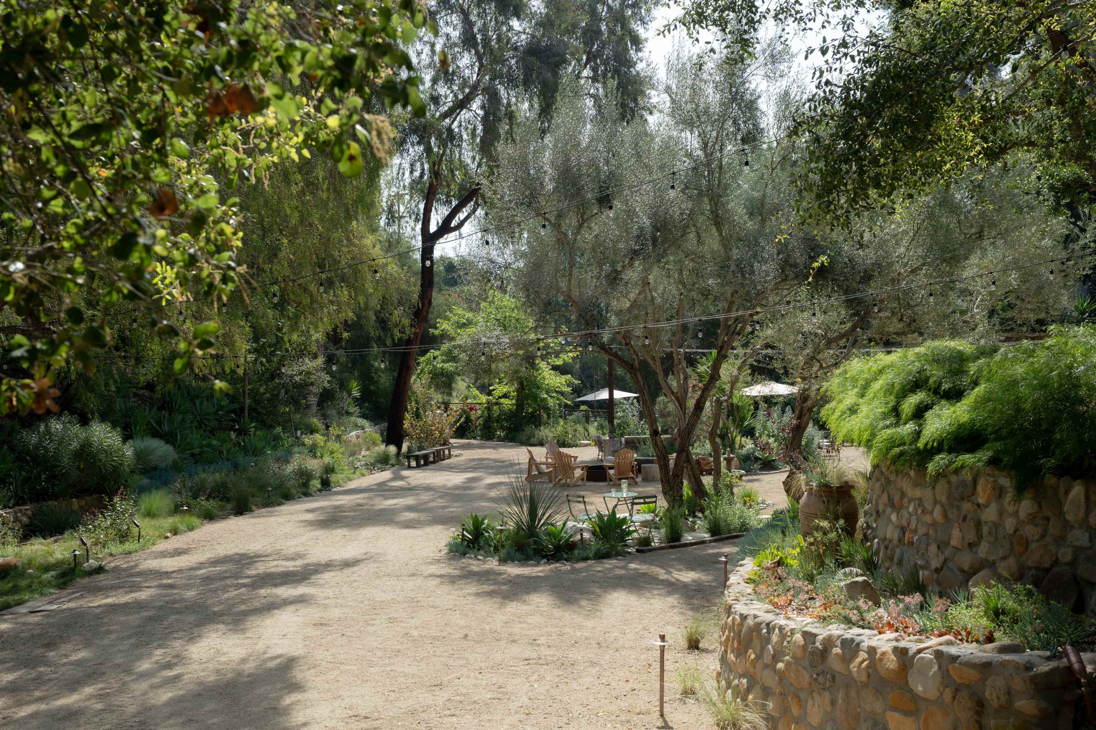 The image shows a sunlit, winding dirt path through a landscaped garden with greenery, stone walls, and outdoor seating areas.