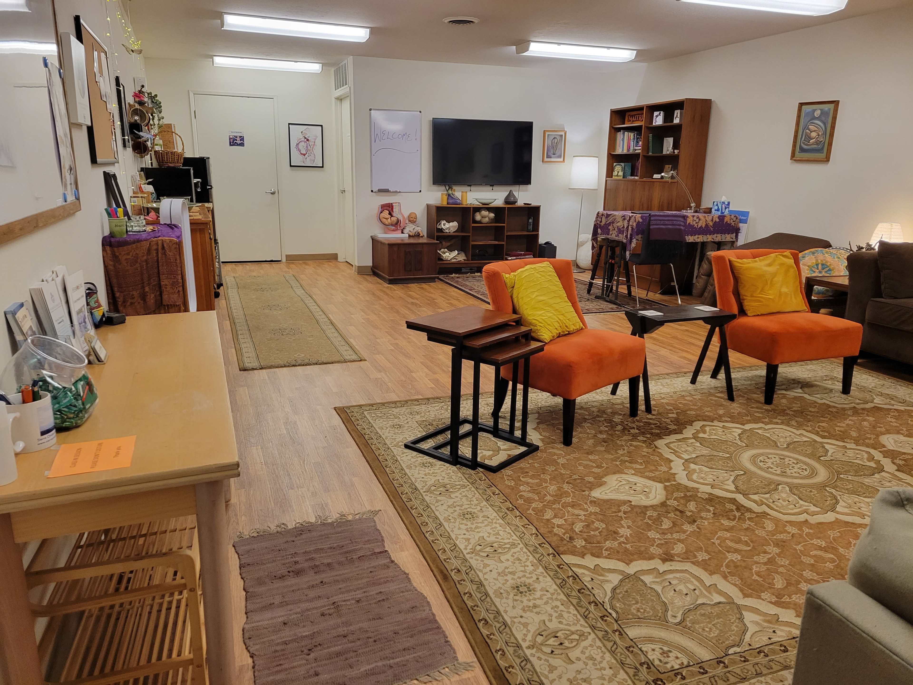 A cozy interior space features orange chairs, a wooden table, shelves, and a television, with rugs on the floor and decorative artwork on the walls.