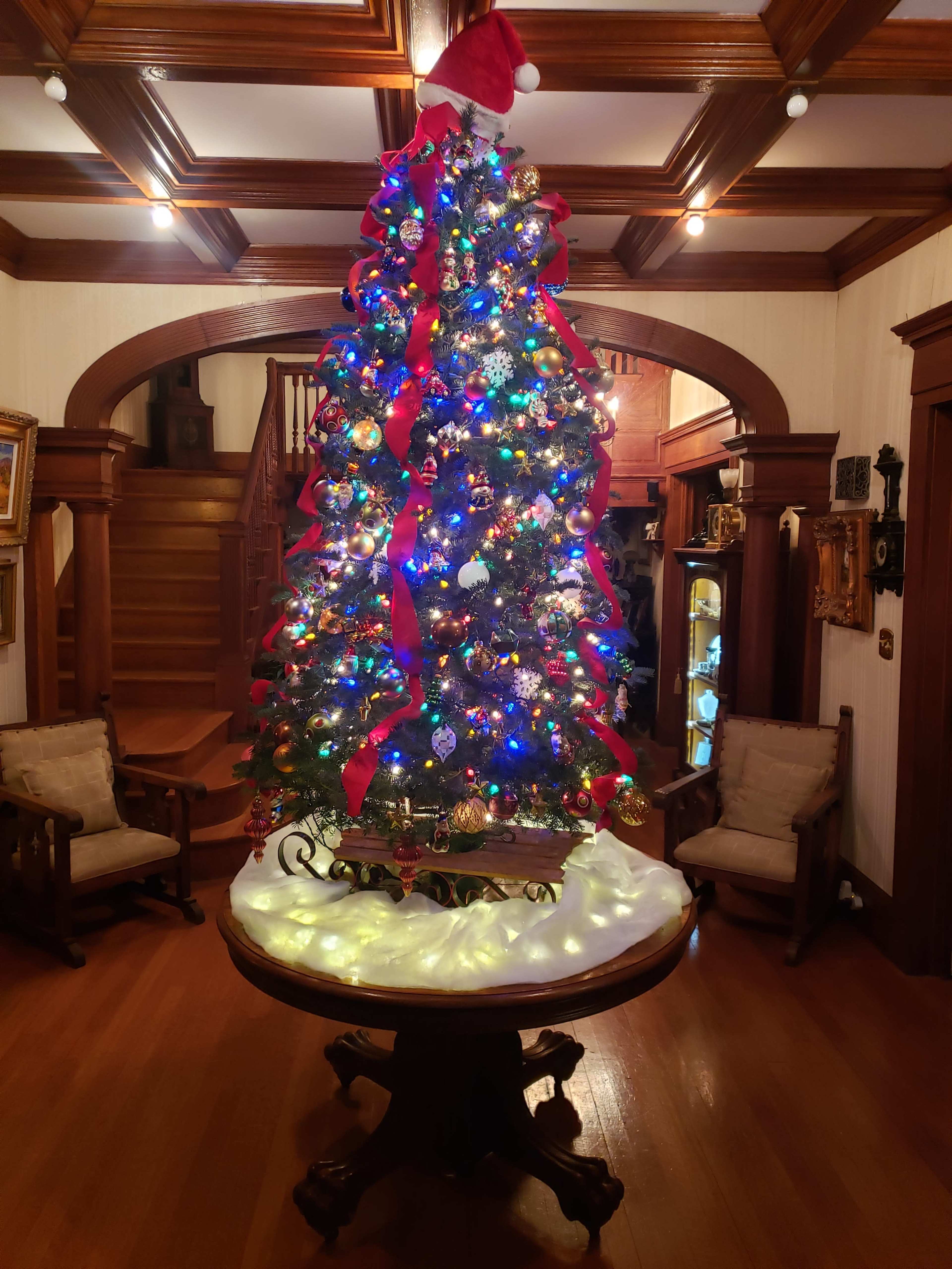A brightly decorated Christmas tree stands on a circular table adorned with white lights in a warmly lit foyer.
