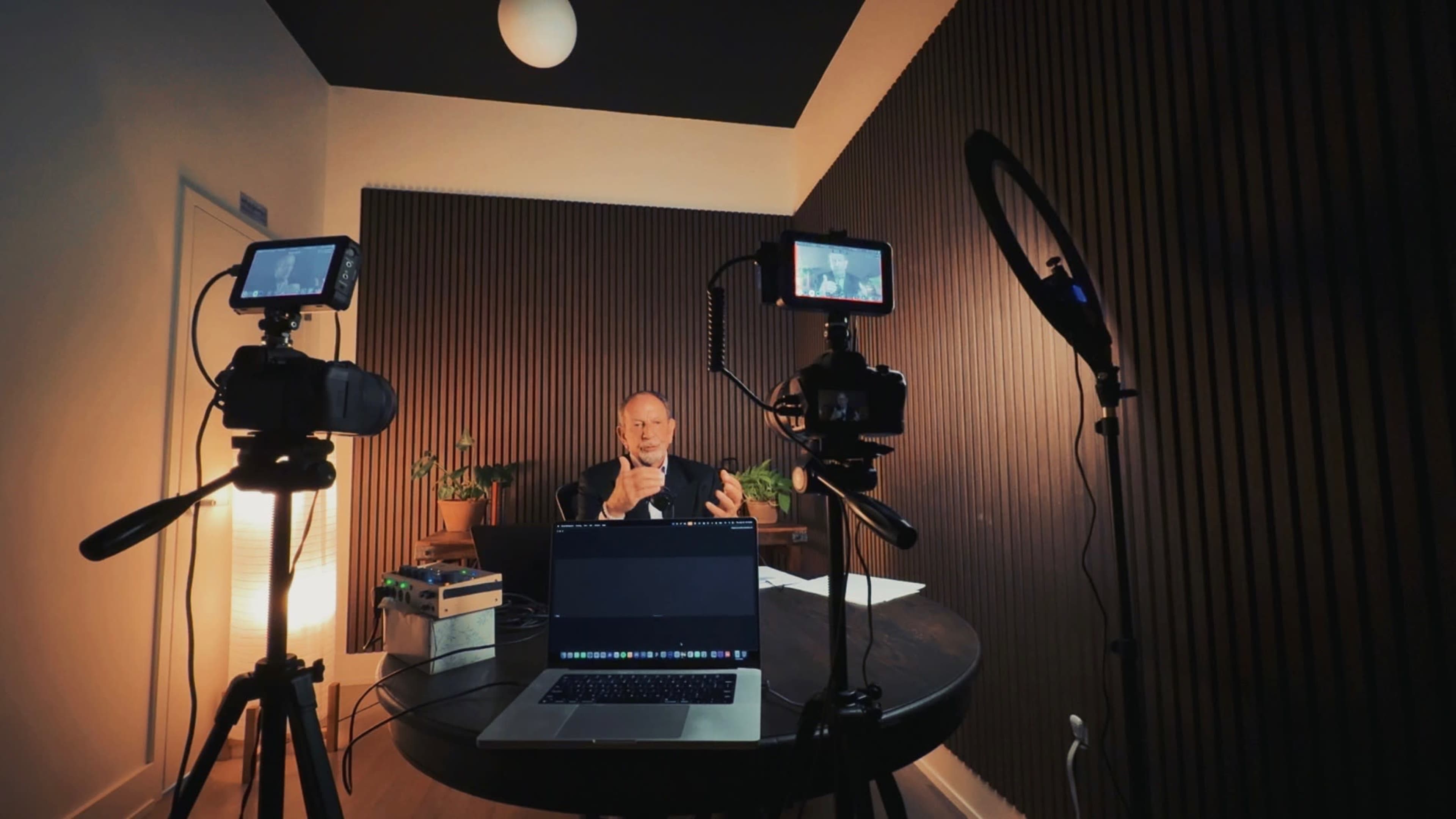 A man is sitting at a table in a dimly lit room, speaking into a microphone while being recorded by cameras on tripods.