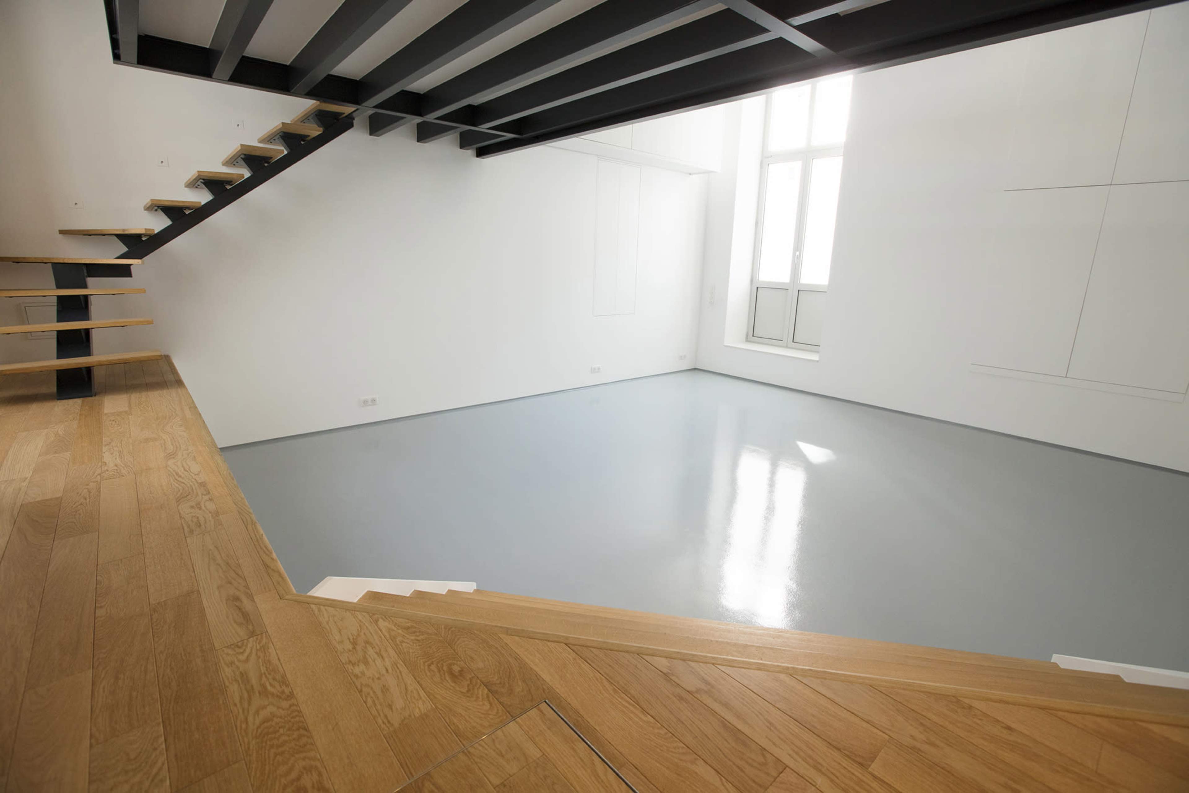 An empty room features a wooden staircase leading to a second level, with large windows allowing natural light to enter.