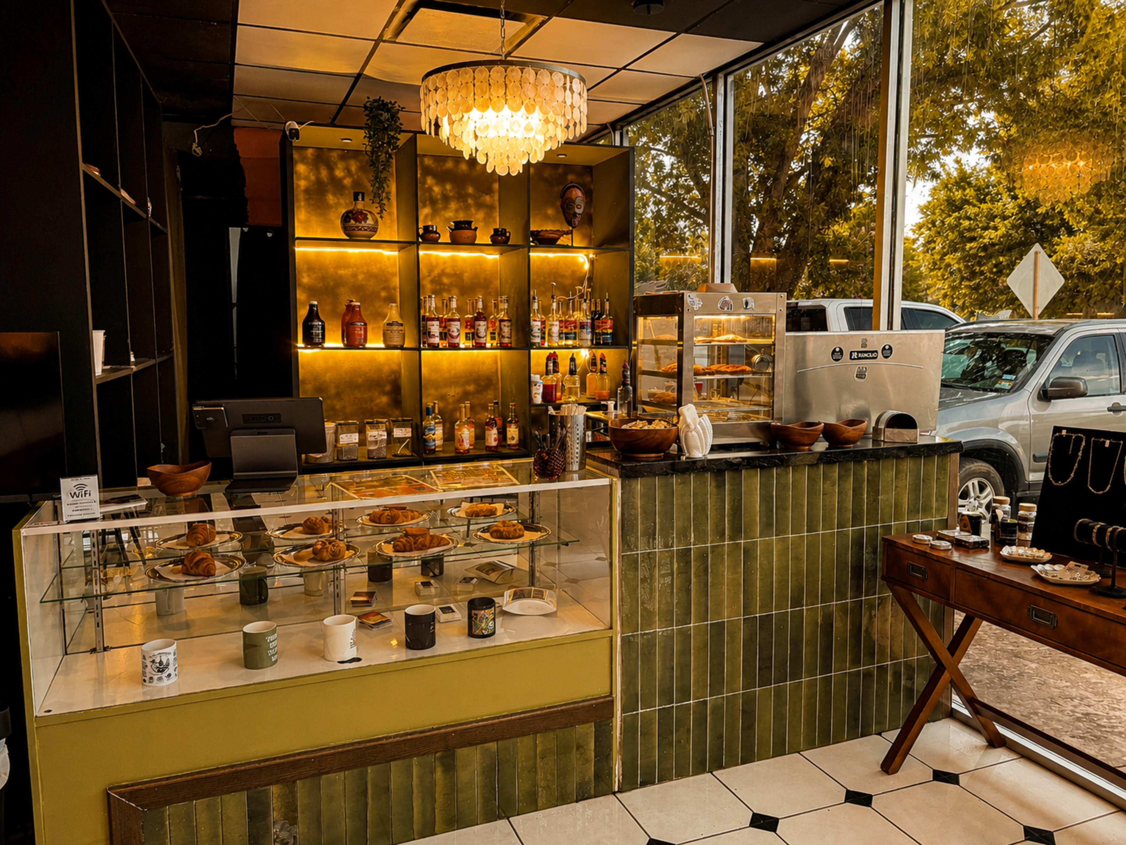 The image shows a small café with a display case filled with baked goods, a counter featuring beverages and kitchen supplies, and large windows providing natural light.
