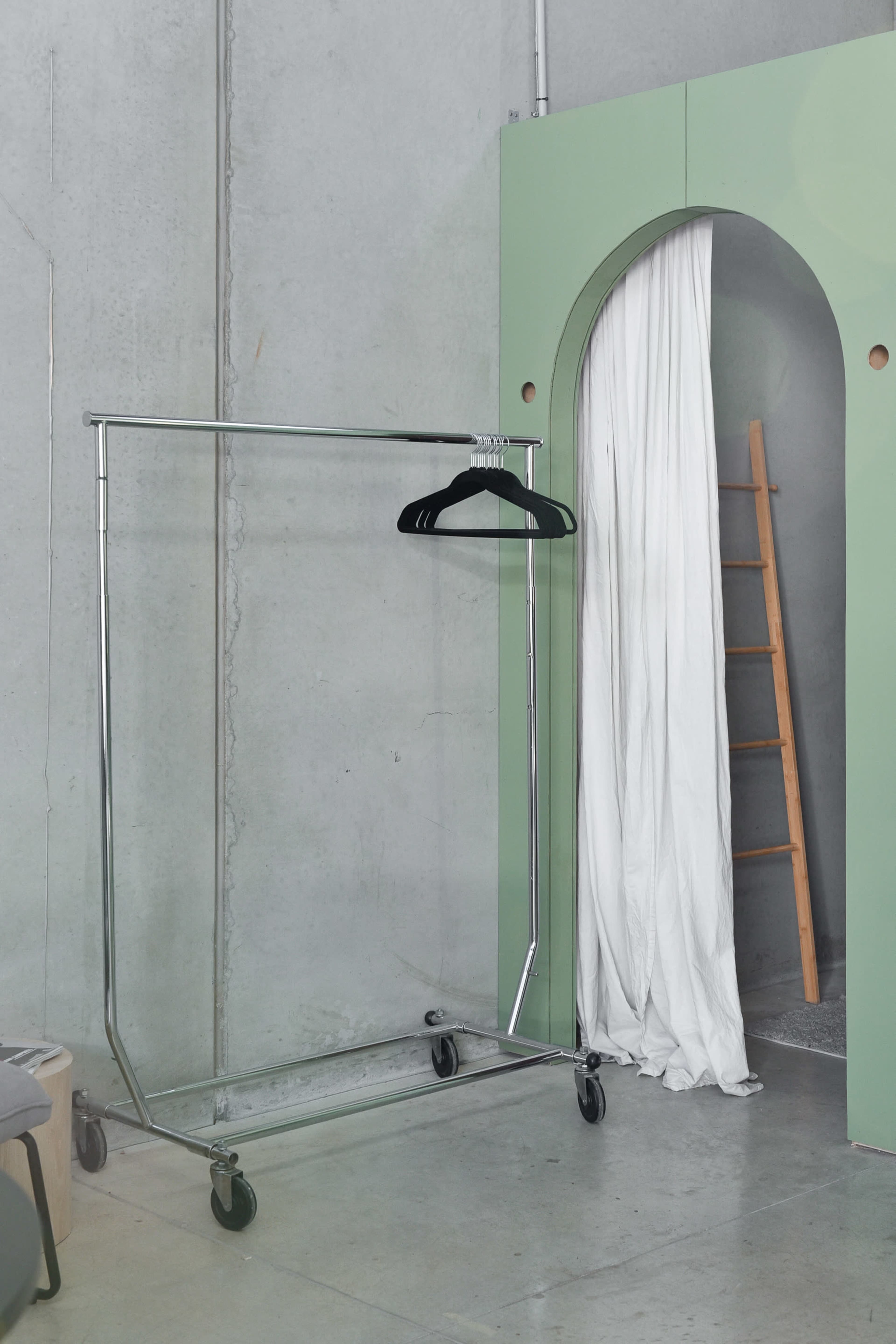 A metal clothing rack on wheels stands next to an arched doorway with a white curtain and a wooden ladder leaning against the wall.