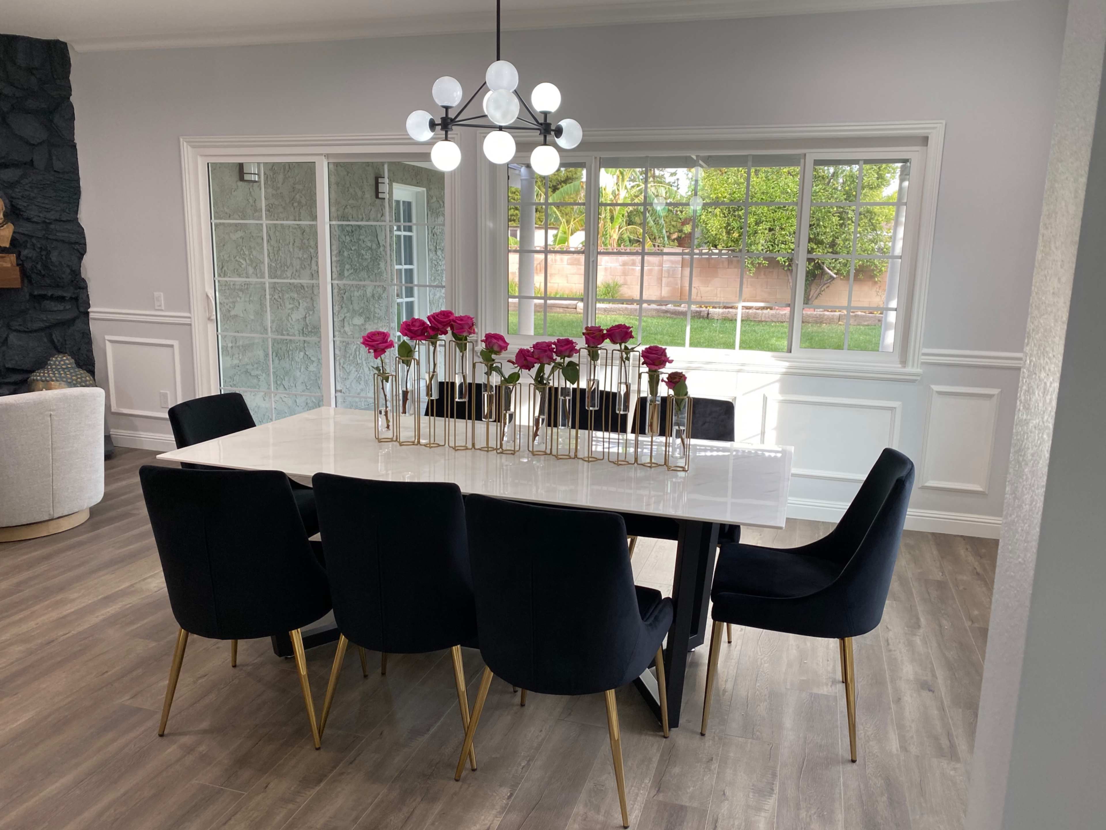 A modern dining room features a large white table surrounded by black chairs, with a vase of pink flowers at the center and sunlight streaming through large windows.