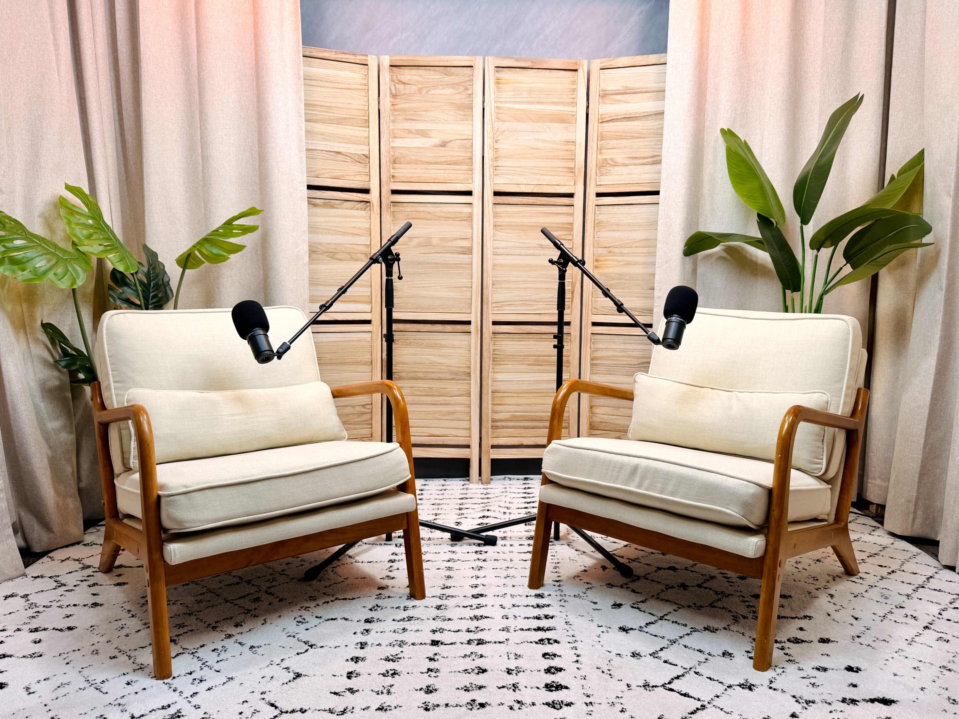 Two beige armchairs with wooden frames are positioned facing each other, accompanied by microphones on stands, in front of a wooden folding screen and decorative plants.
