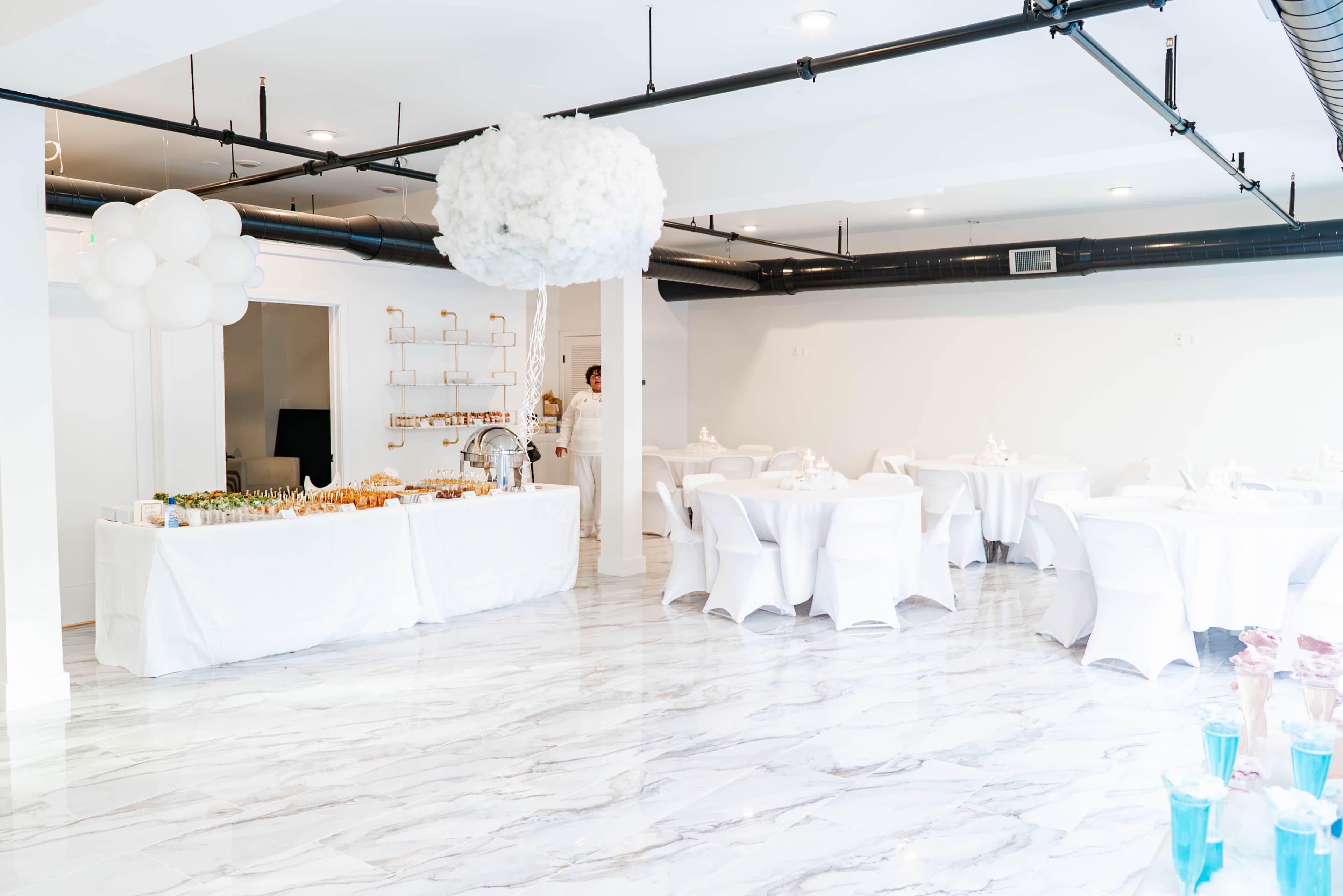 A brightly lit banquet room with round tables covered in white cloth and a buffet table displaying various food items against a marble floor.