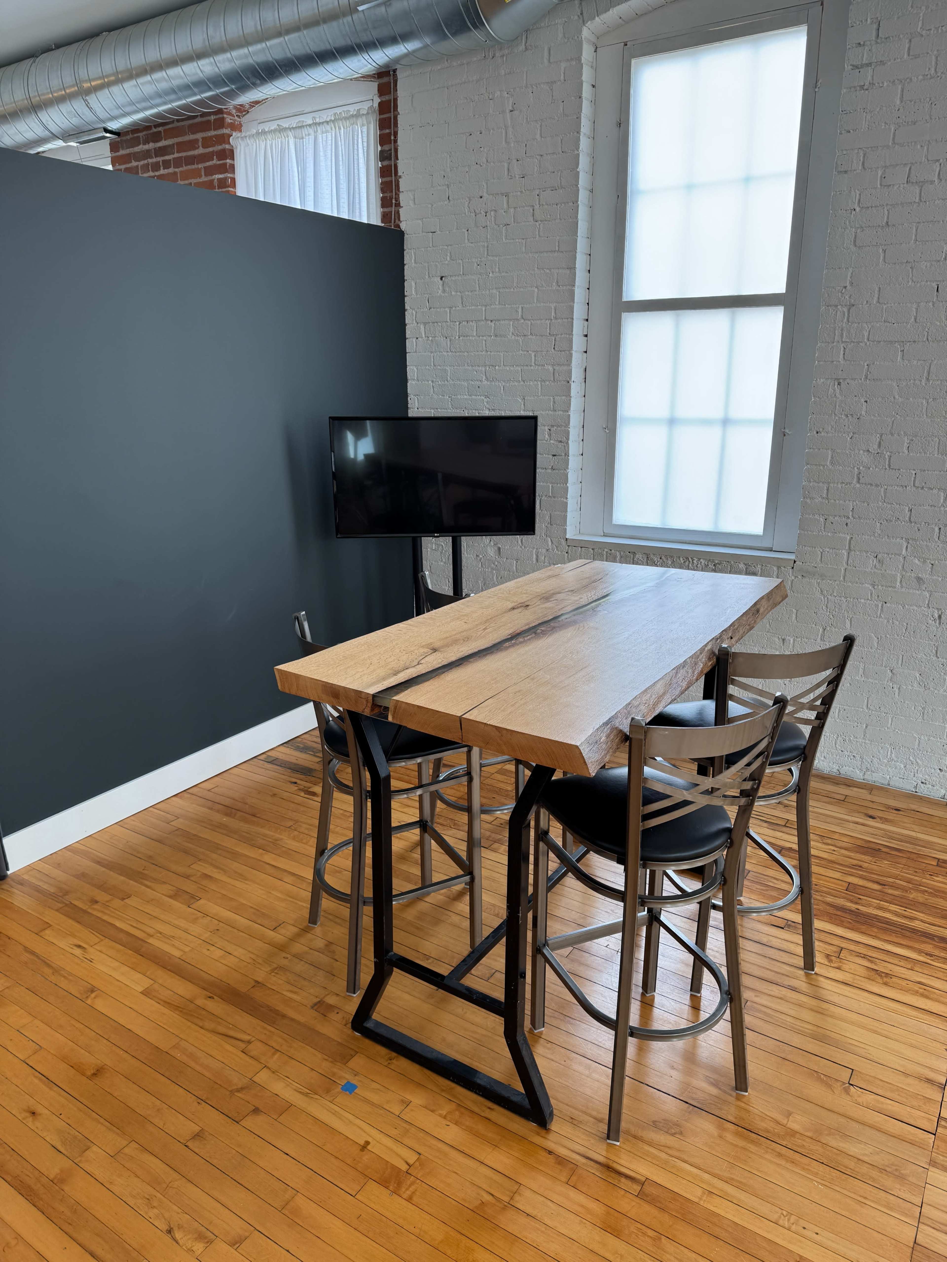 A wooden table with a live edge is positioned in a room with hardwood floors, accompanied by four black chairs and a wall-mounted television.