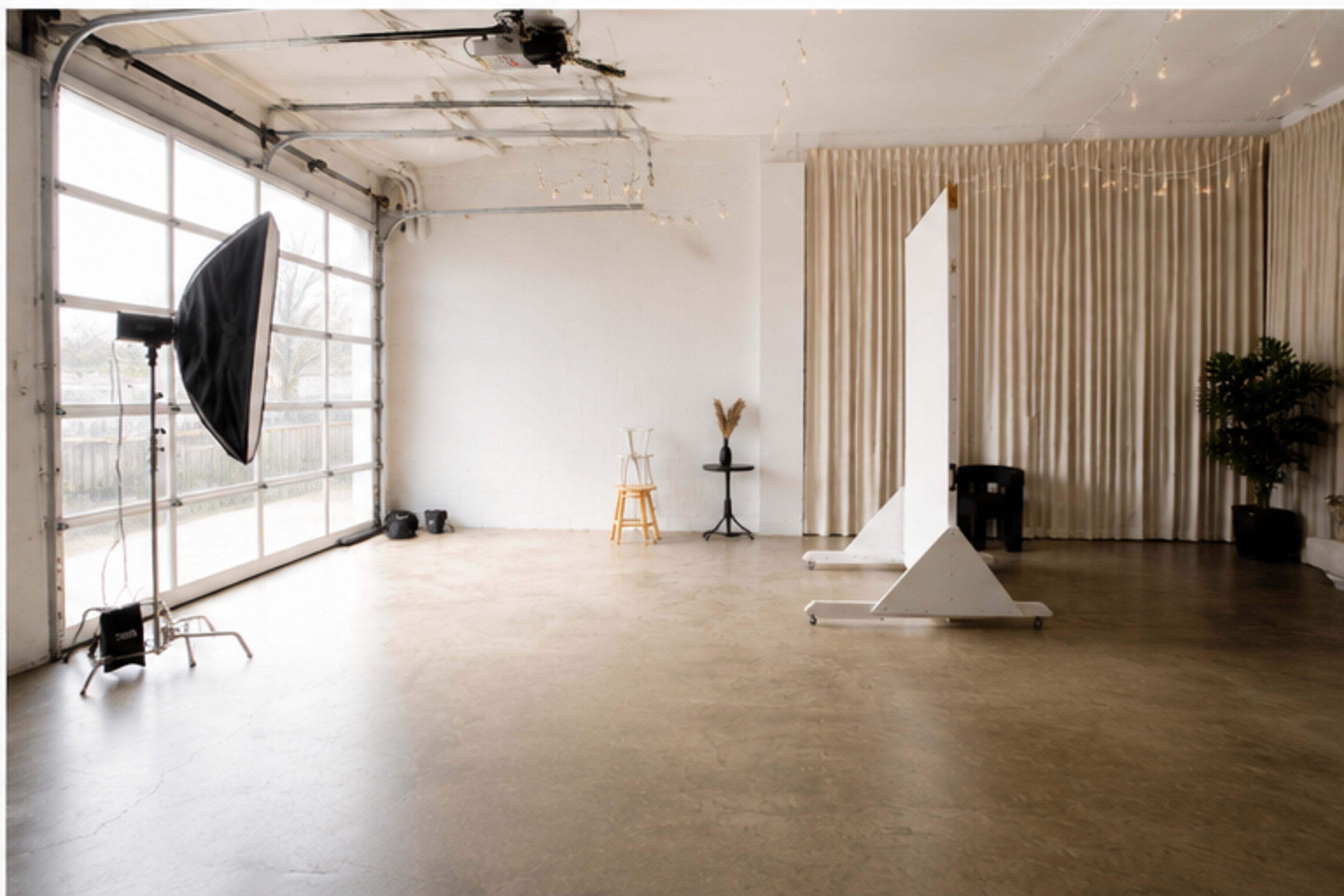 The image shows a well-lit studio space featuring a large window, a black photography umbrella, two stools, a tall plant, and a backdrop draped in fabric.