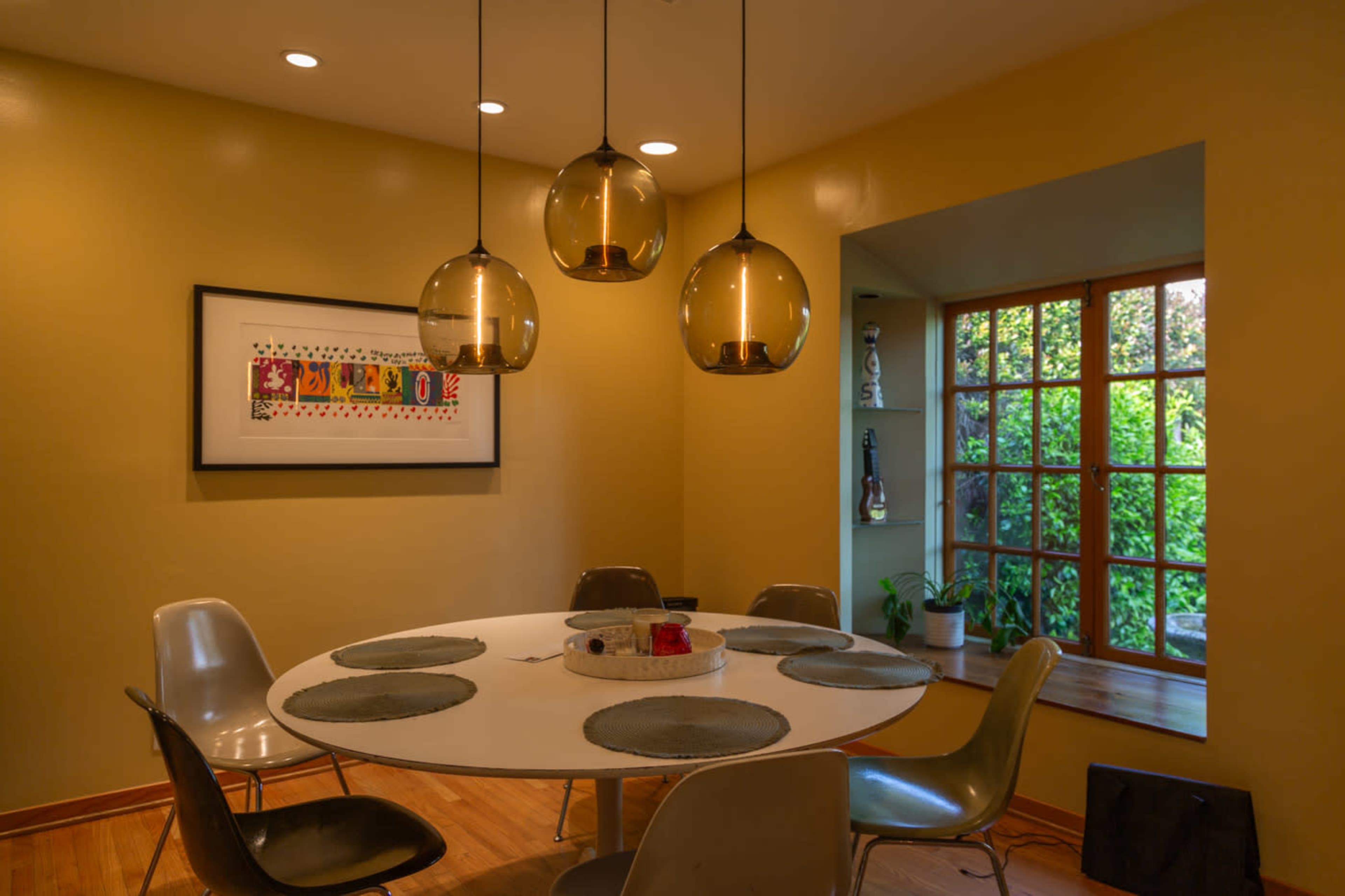A round dining table surrounded by chairs sits beneath three pendant lights in a room with yellow walls and a window showcasing greenery outside.