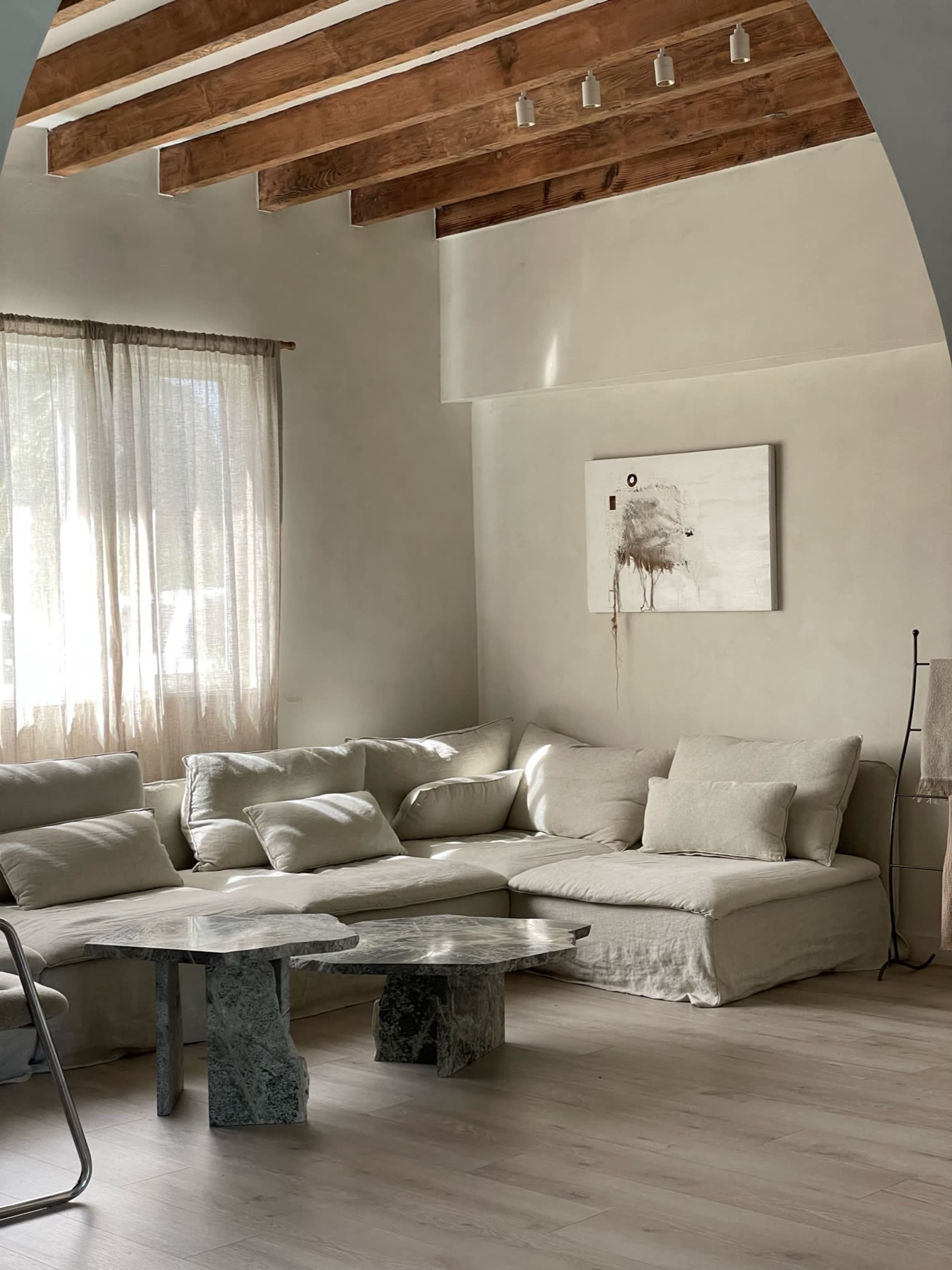 A light-filled living room features a large, sectional sofa and a unique coffee table made of stone on a wooden floor, with exposed wooden beams overhead.