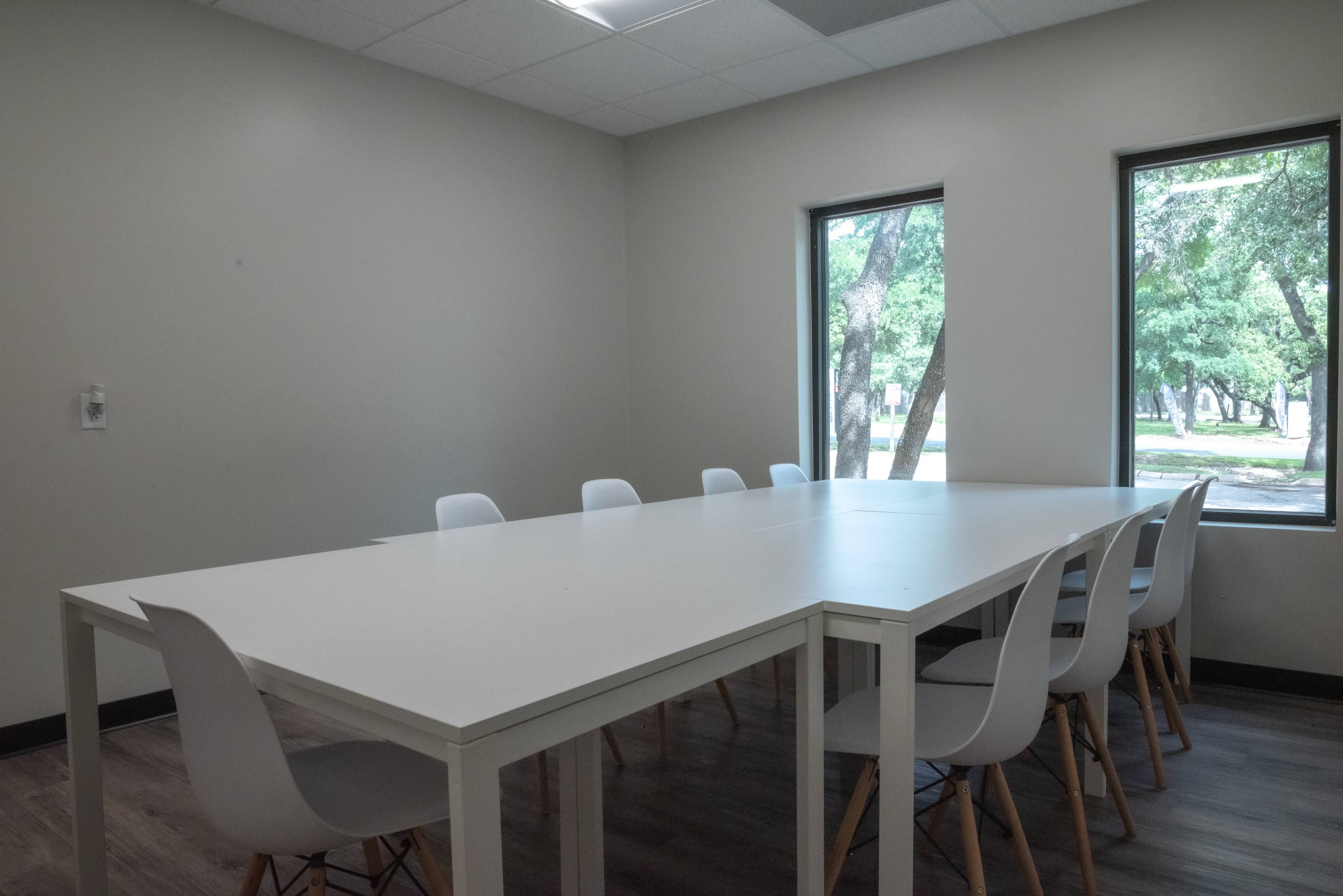 A spacious meeting room features a large white table surrounded by several chairs and is illuminated by natural light from two windows.