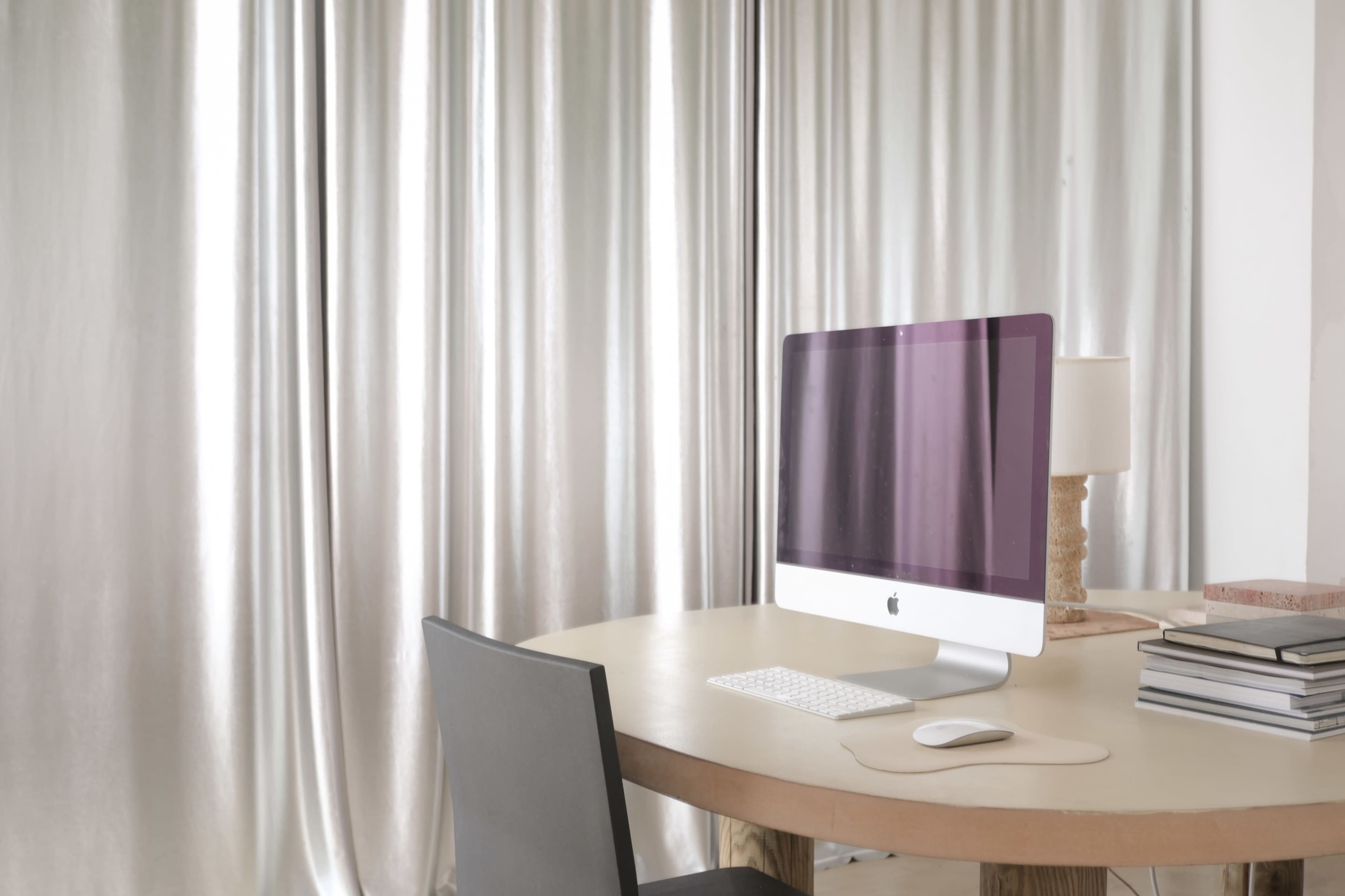 A modern workspace features an iMac on a round table next to a lamp and stacked books, with sheer curtains in the background.