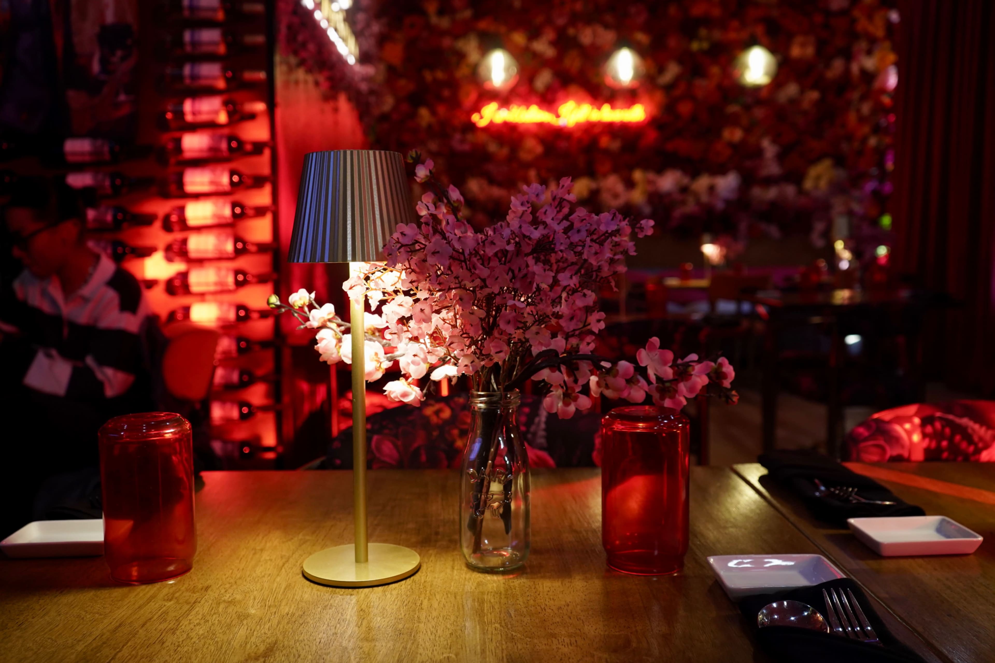 A table set in a restaurant features a vase of pink flowers and a lamp, with a backdrop of wine bottles and red lighting.