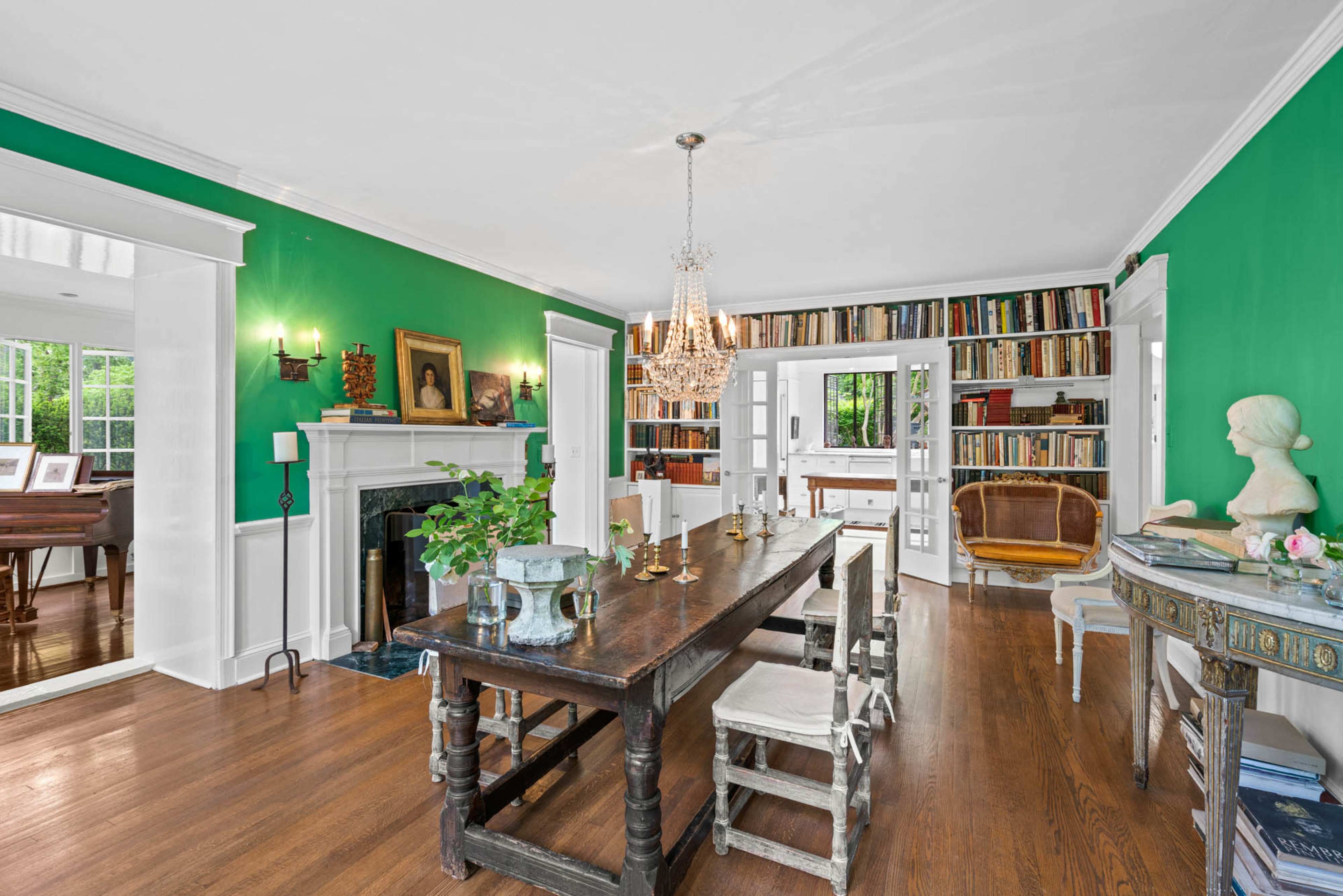 The image shows a spacious dining area with a long wooden table, surrounded by chairs, a chandelier overhead, and walls painted in green, along with a bookshelf and decorative items.