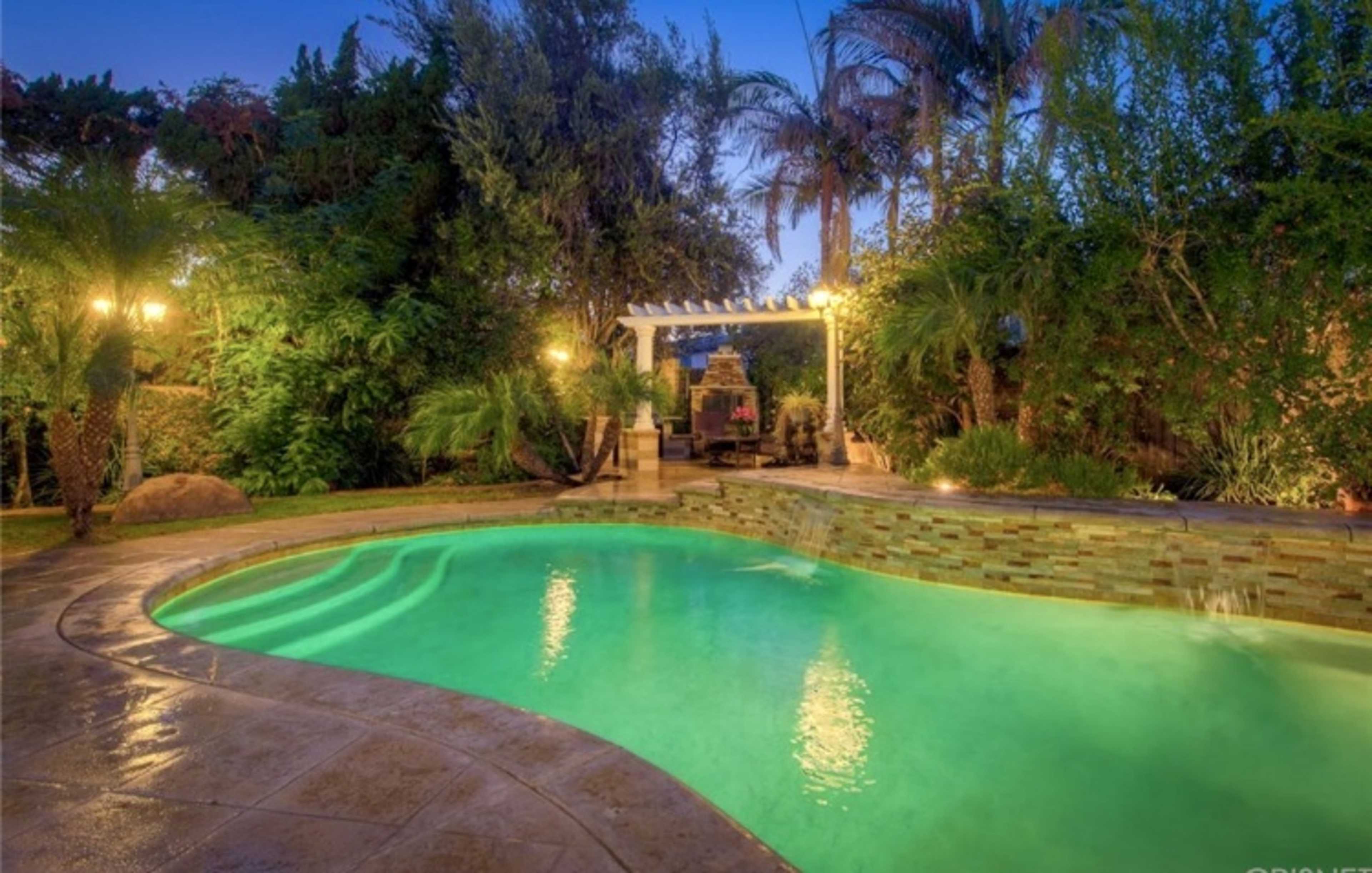 The image shows a well-lit backyard featuring a swimming pool surrounded by lush greenery and a seating area under a pergola.