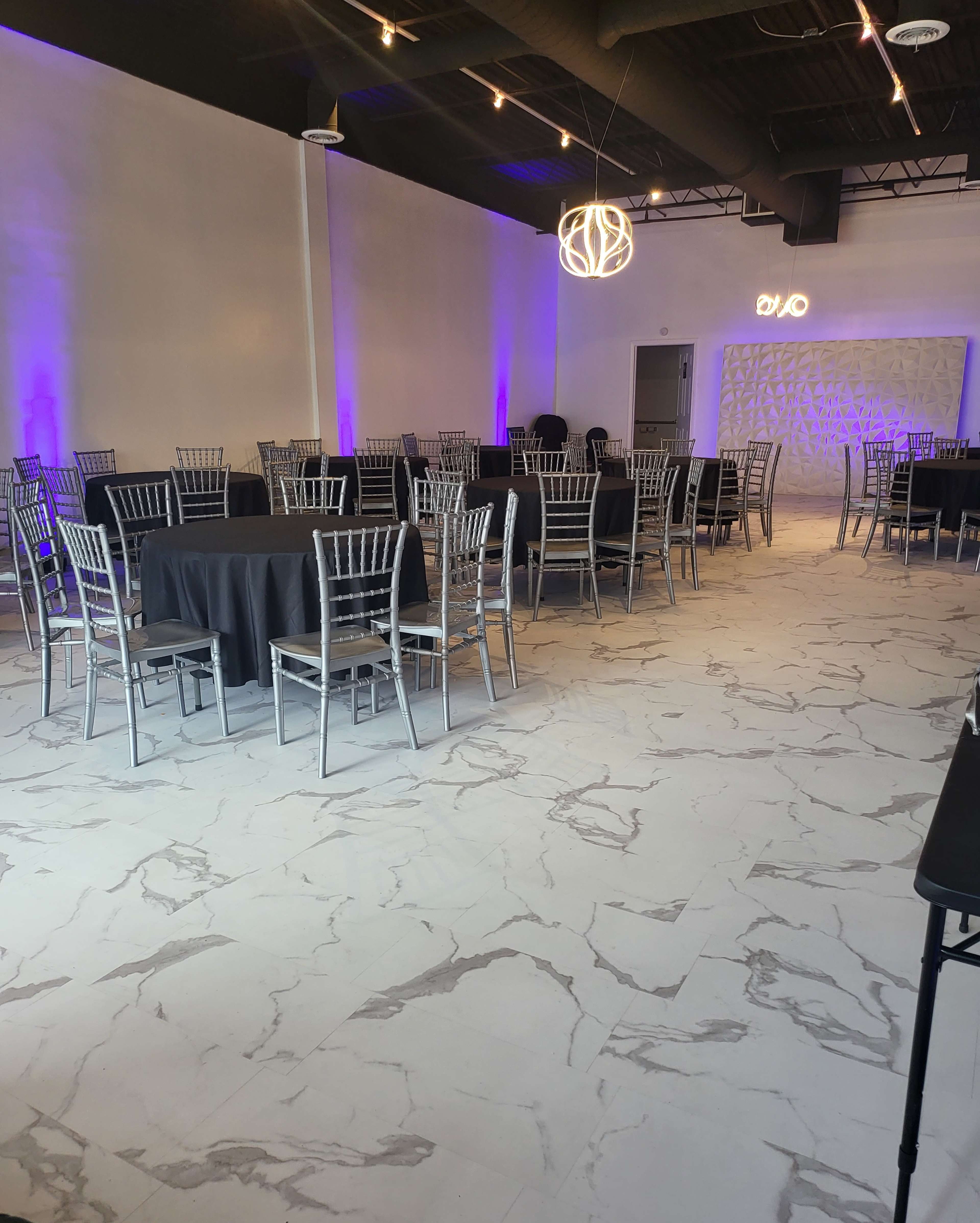 A spacious event venue featuring several round tables covered with black tablecloths, silver chairs, and purple ambient lighting.
