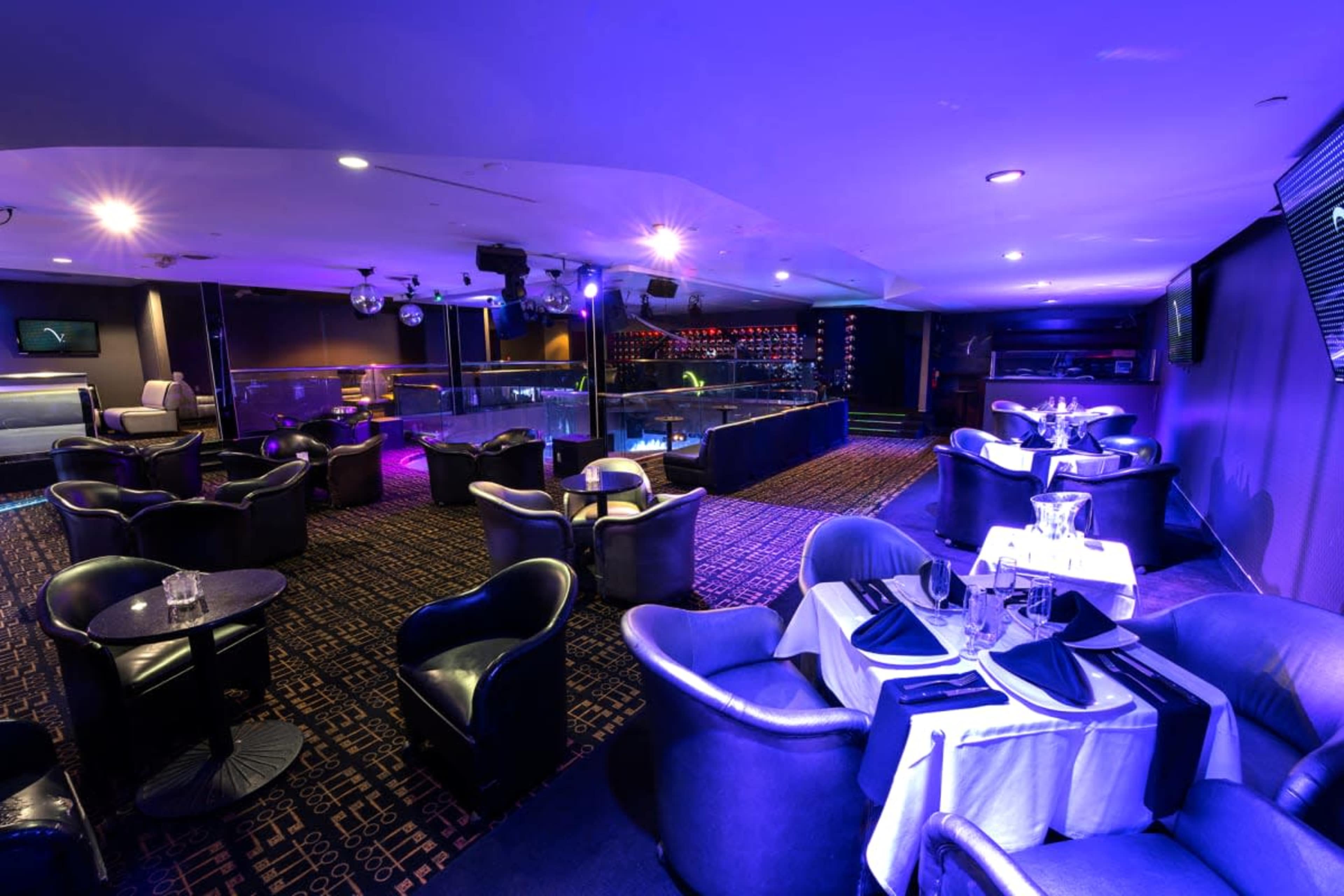 The image shows a dimly lit lounge with stylish furniture, including round tables and comfortable chairs, and a bar area illuminated by colorful lights.