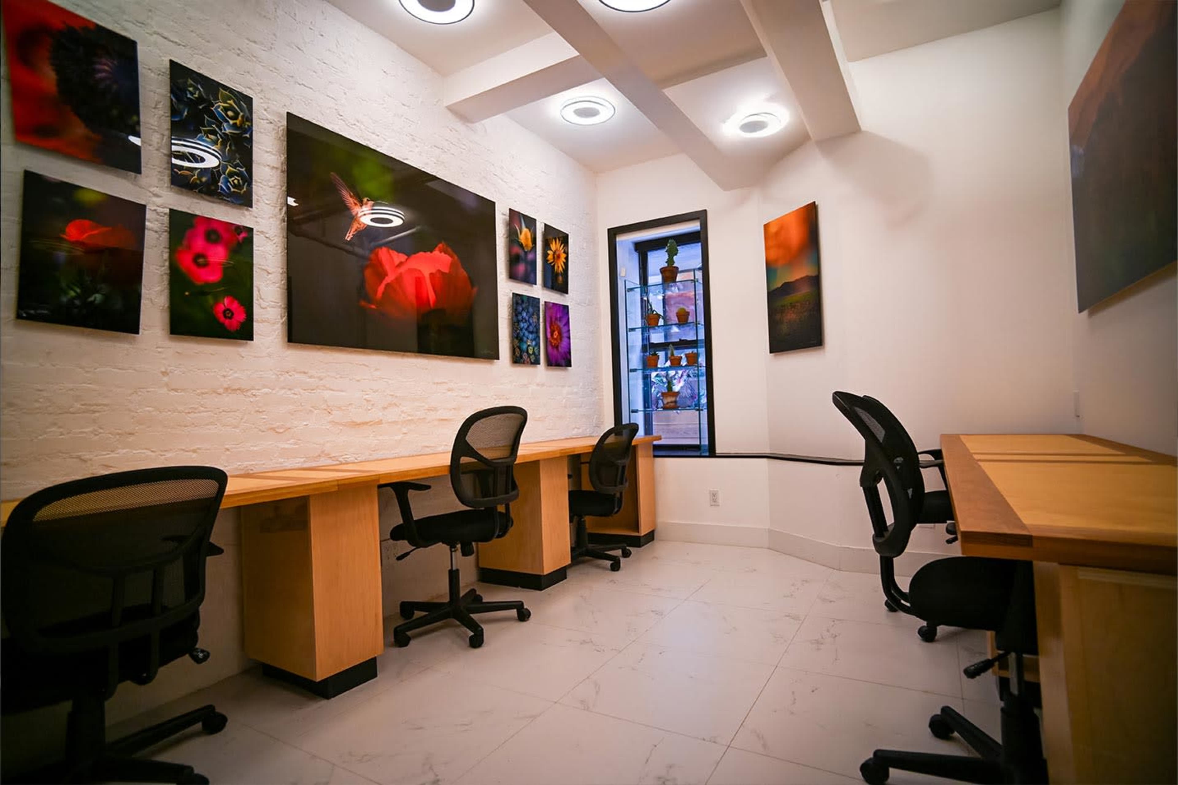 The image shows a modern workspace with two wooden desks and ergonomic chairs, surrounded by colorful framed artwork on the walls.