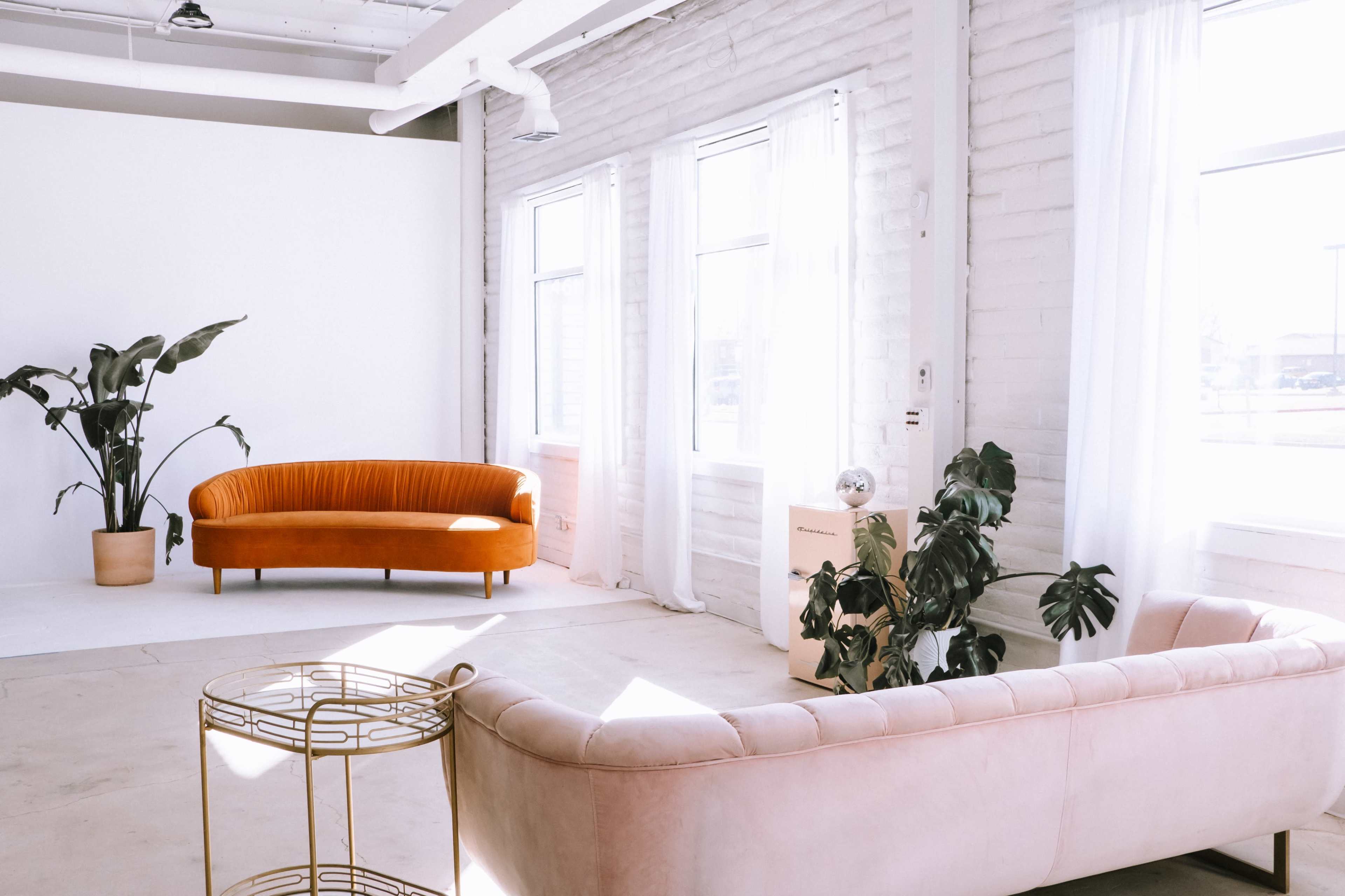 The image shows a bright, minimalist interior featuring an orange curved sofa and a light pink curved sofa, with large windows allowing natural light to fill the space.