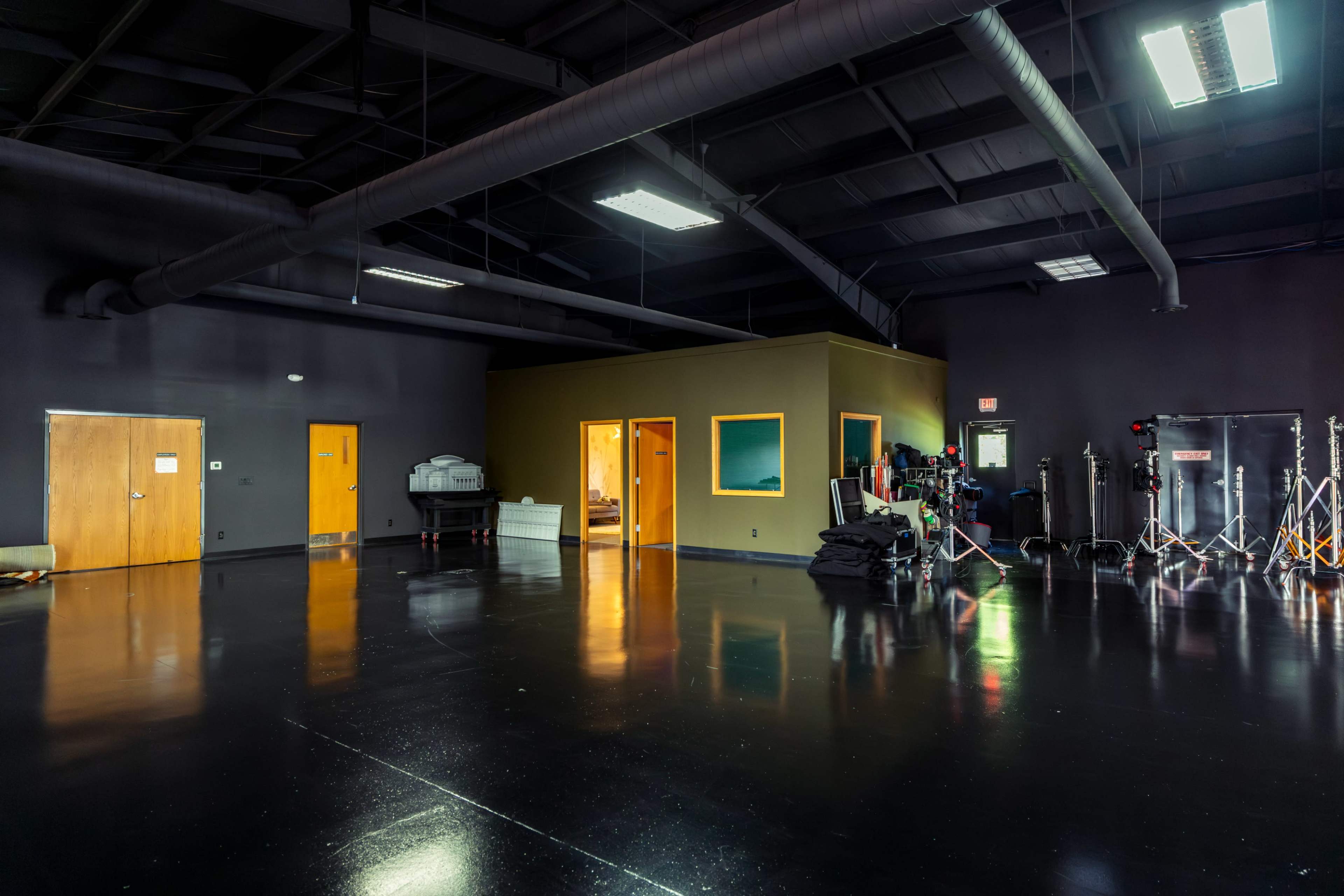 The image shows a large, empty studio space with dark walls, polished floors, and various equipment and rooms along the perimeter.