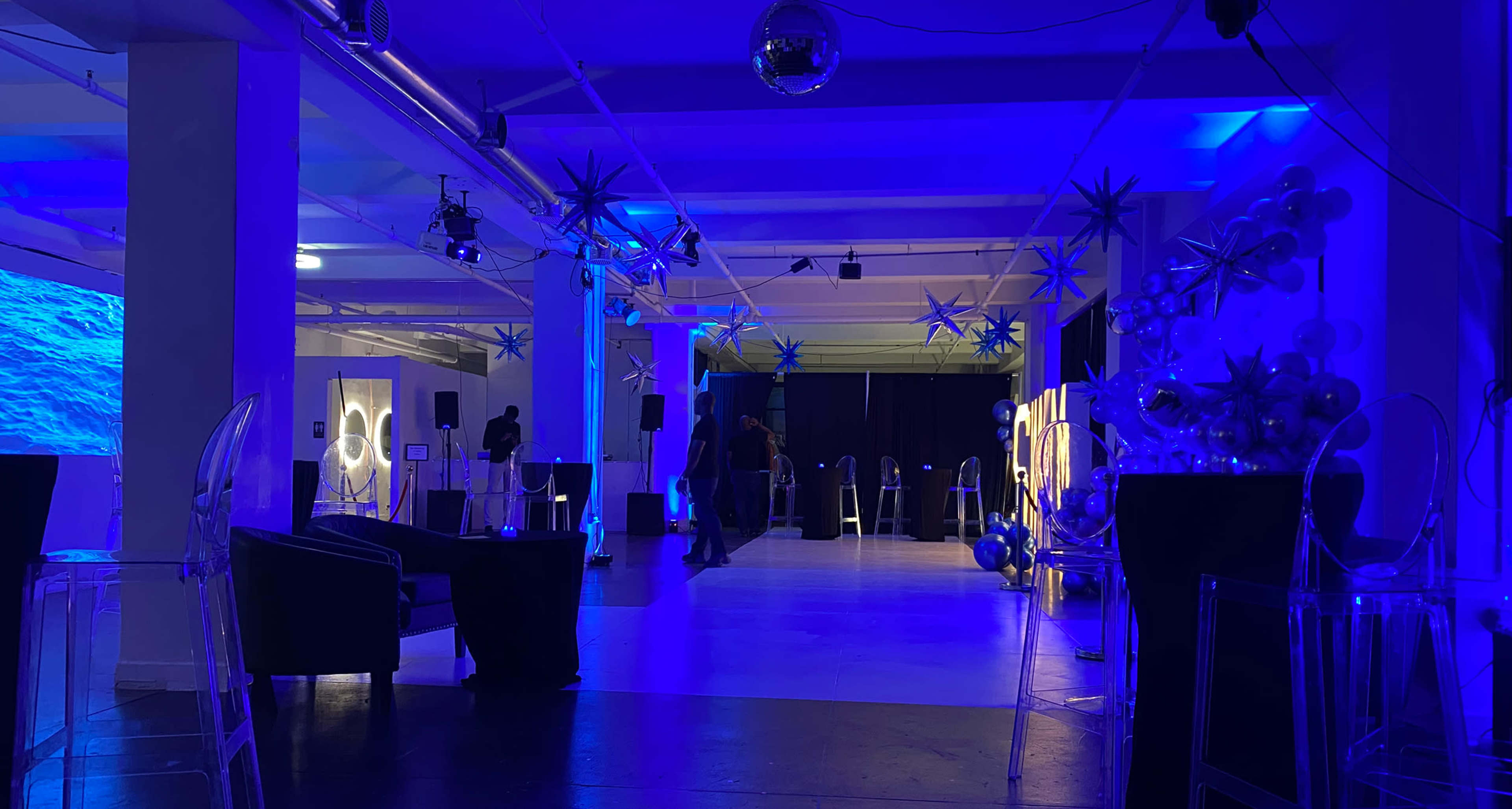 The image shows an event space decorated with blue lighting, featuring chairs, a dance floor, and various decorations.