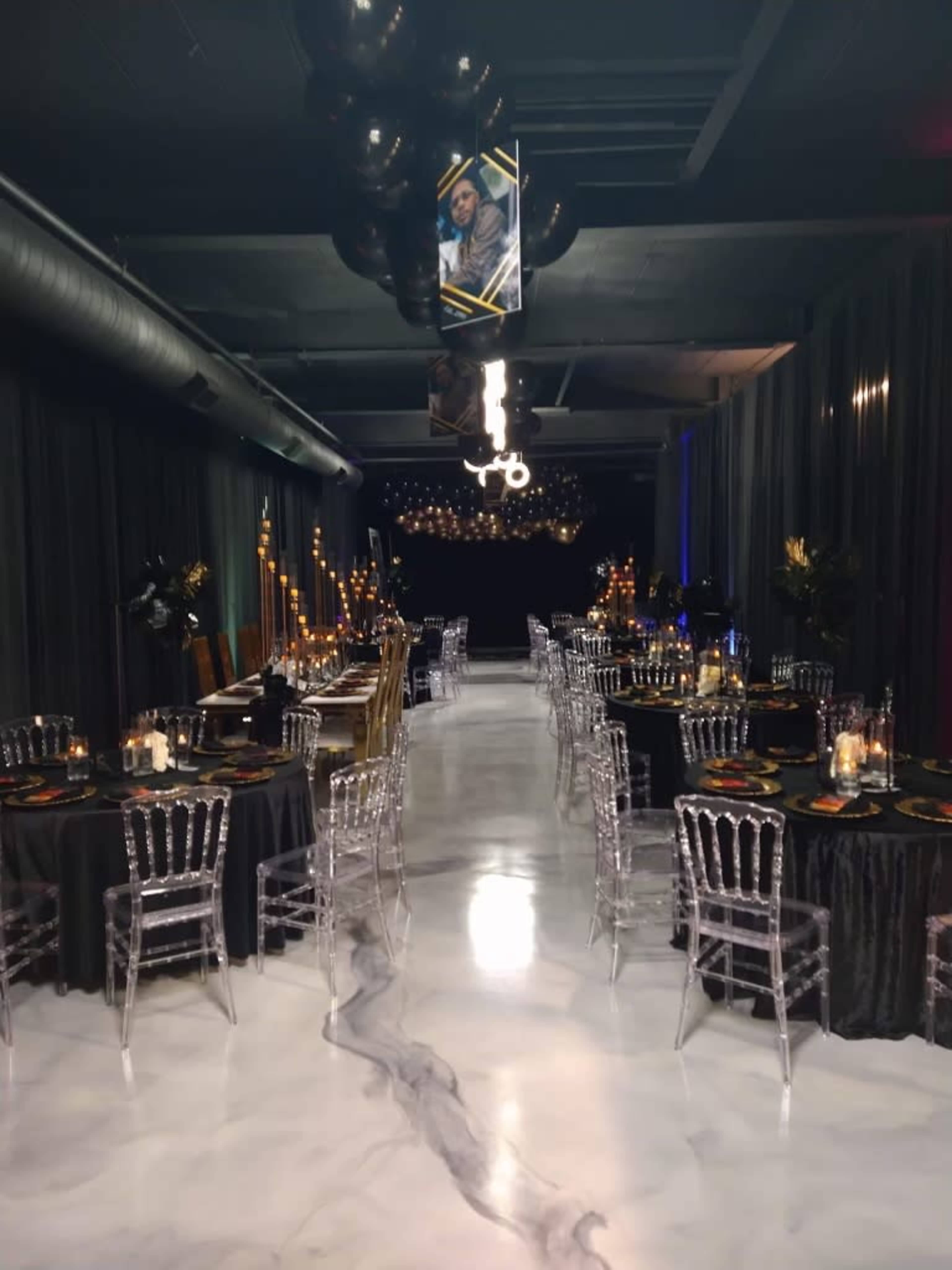 The image shows a dimly lit event space with two rows of elegantly decorated tables set for a formal dinner, featuring black tablecloths and transparent chairs.