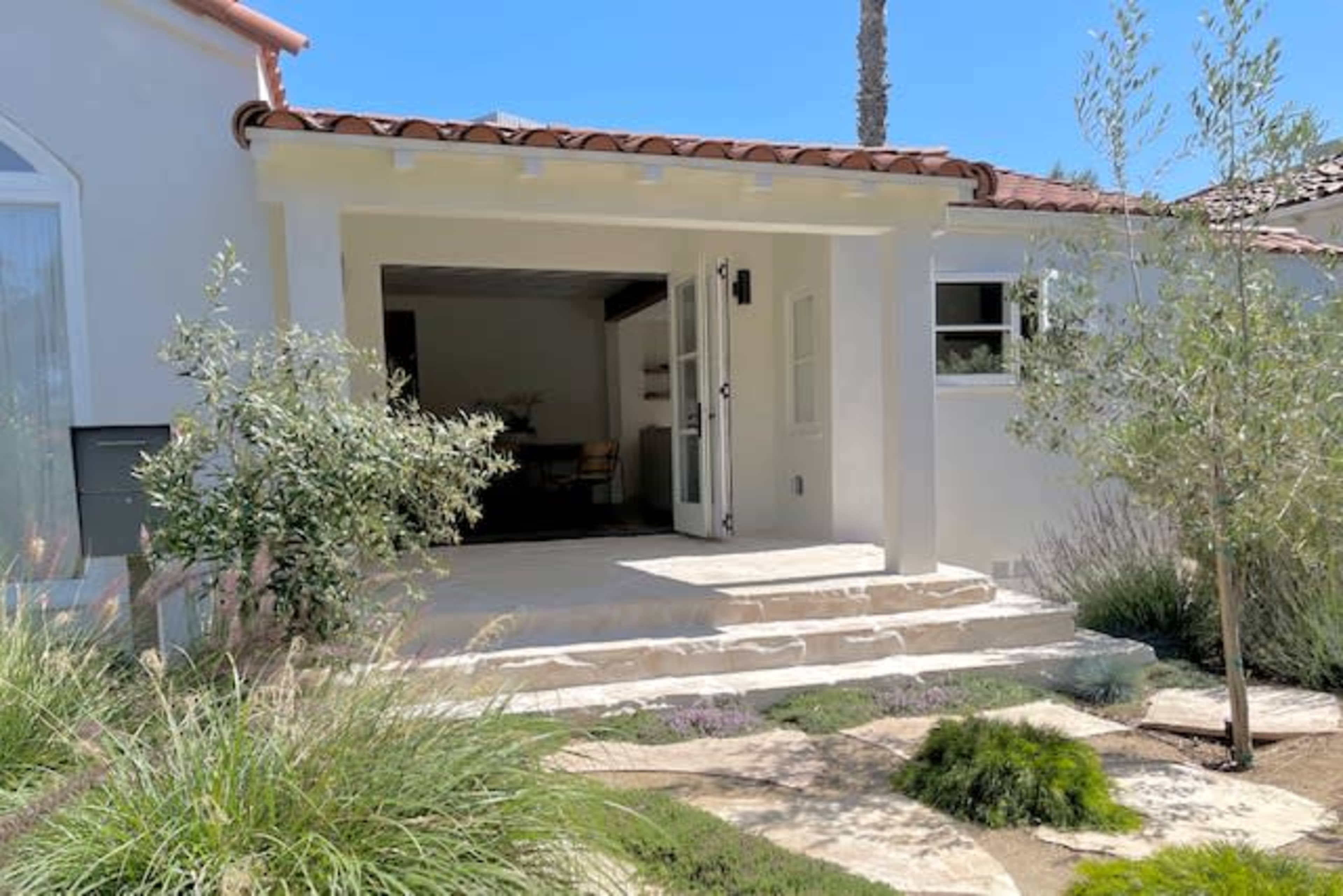 The image shows a white house entrance with a tiled roof, stone steps, and a front porch leading into an open space.
