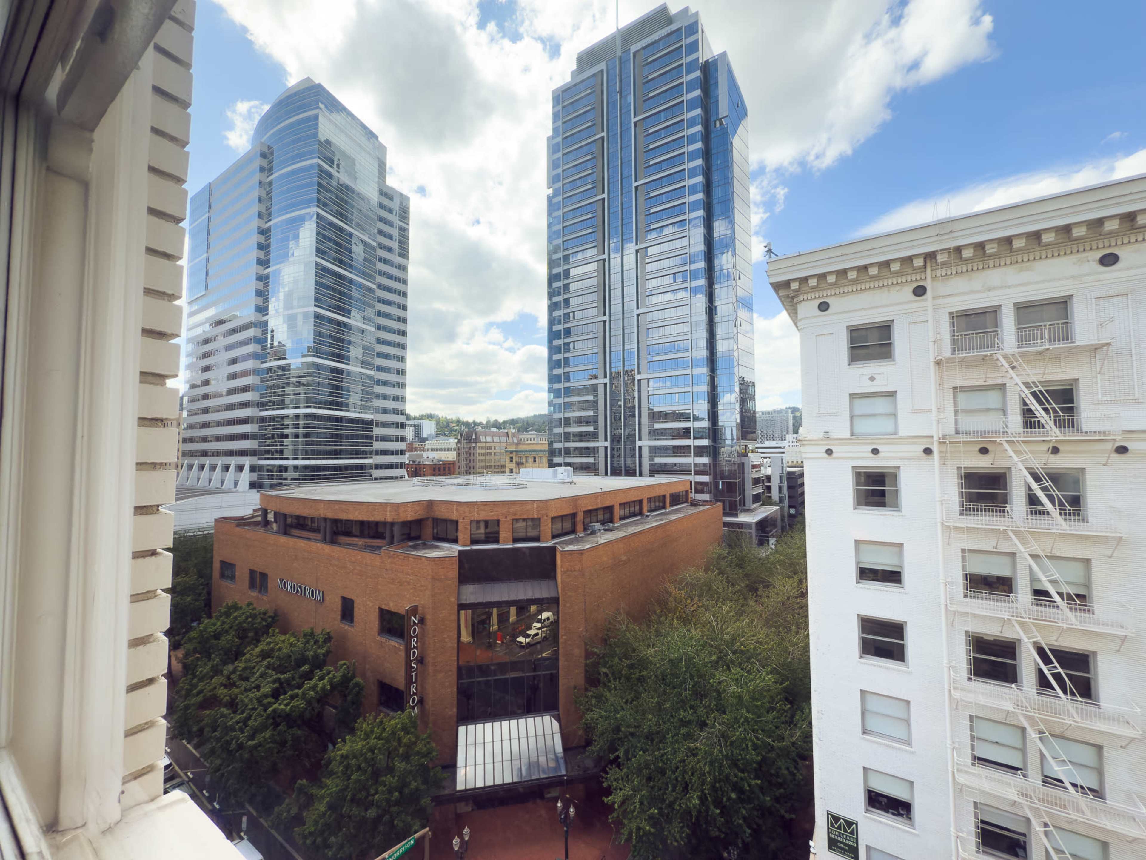 The image shows a view from a window overlooking a cityscape featuring modern skyscrapers and a brick building surrounded by trees.