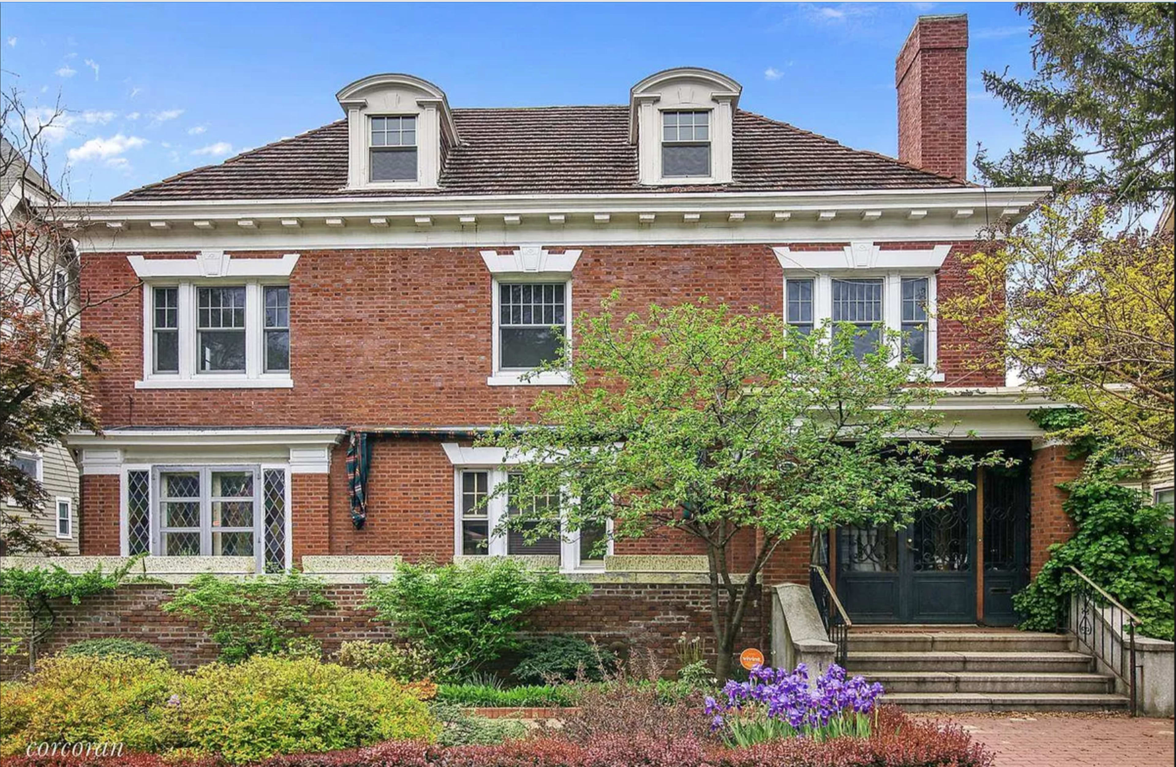 The image shows a large brick house with a symmetrical facade, featuring three dormer windows and a front entrance with a set of stairs surrounded by green shrubs and flowers.
