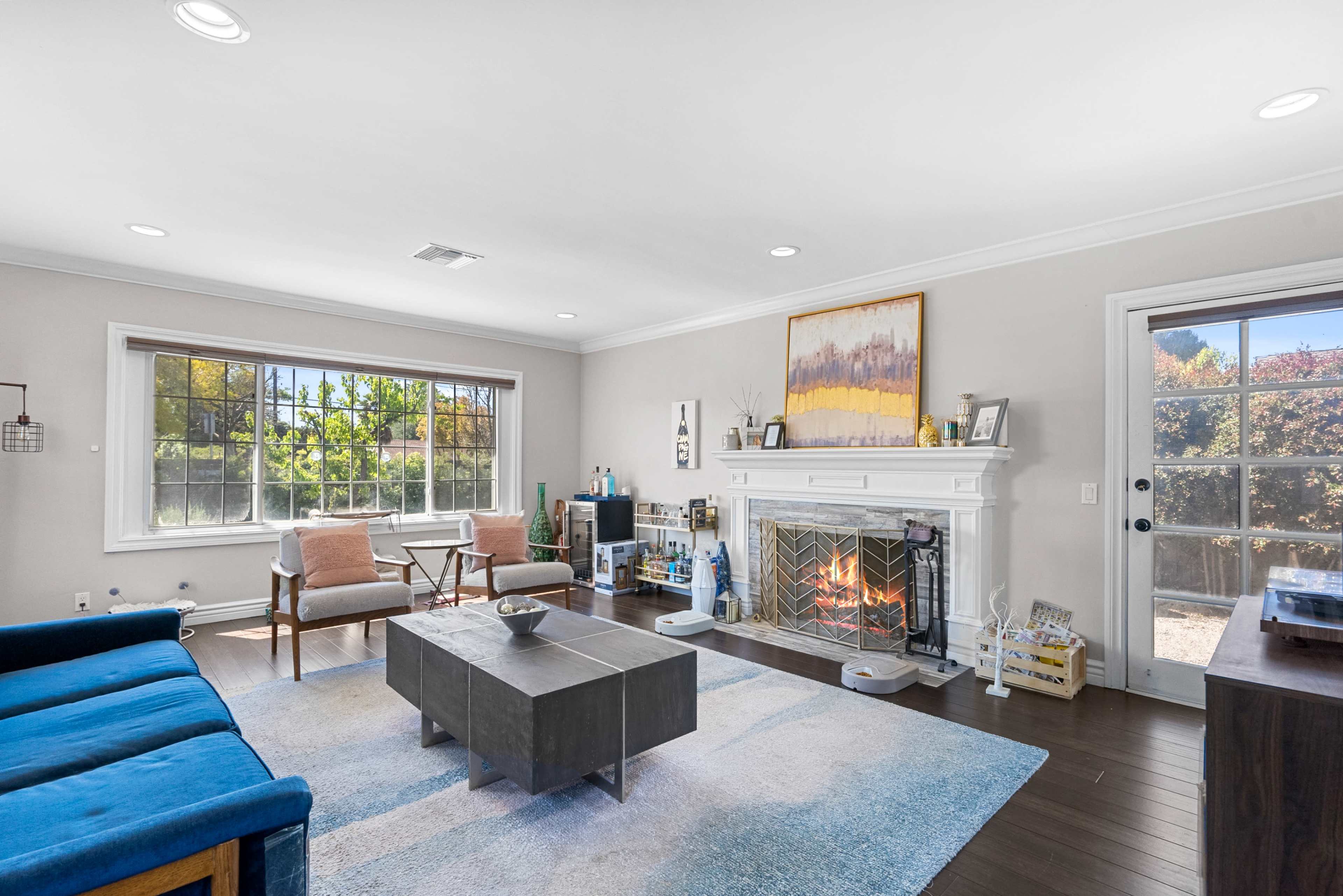 A living room features a blue couch, two armchairs, a coffee table, and a fireplace with a large window and a door leading outside.