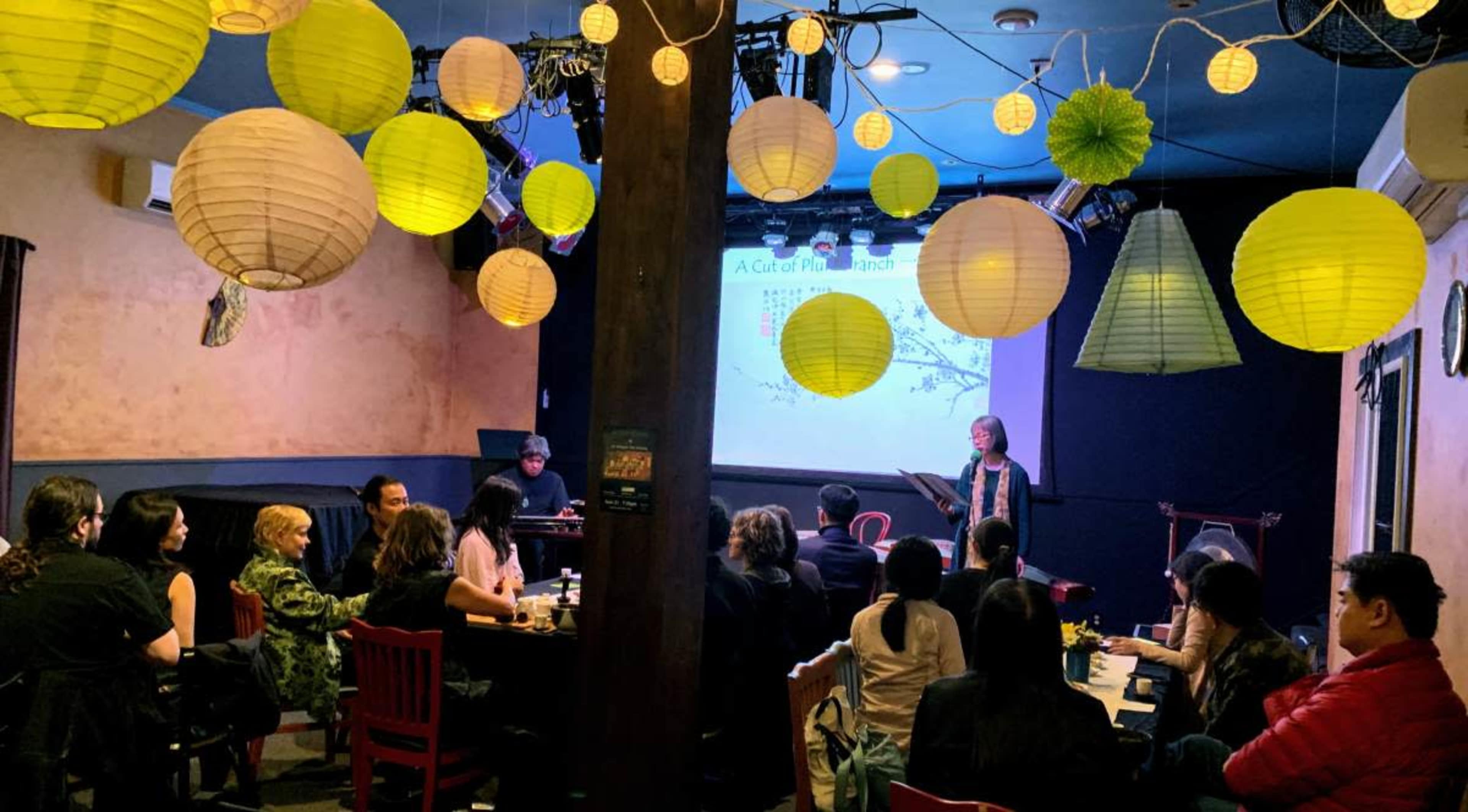 A group of people sits at tables in a dimly lit venue with colorful lanterns hanging from the ceiling while a presenter speaks in front of a screen displaying visuals.
