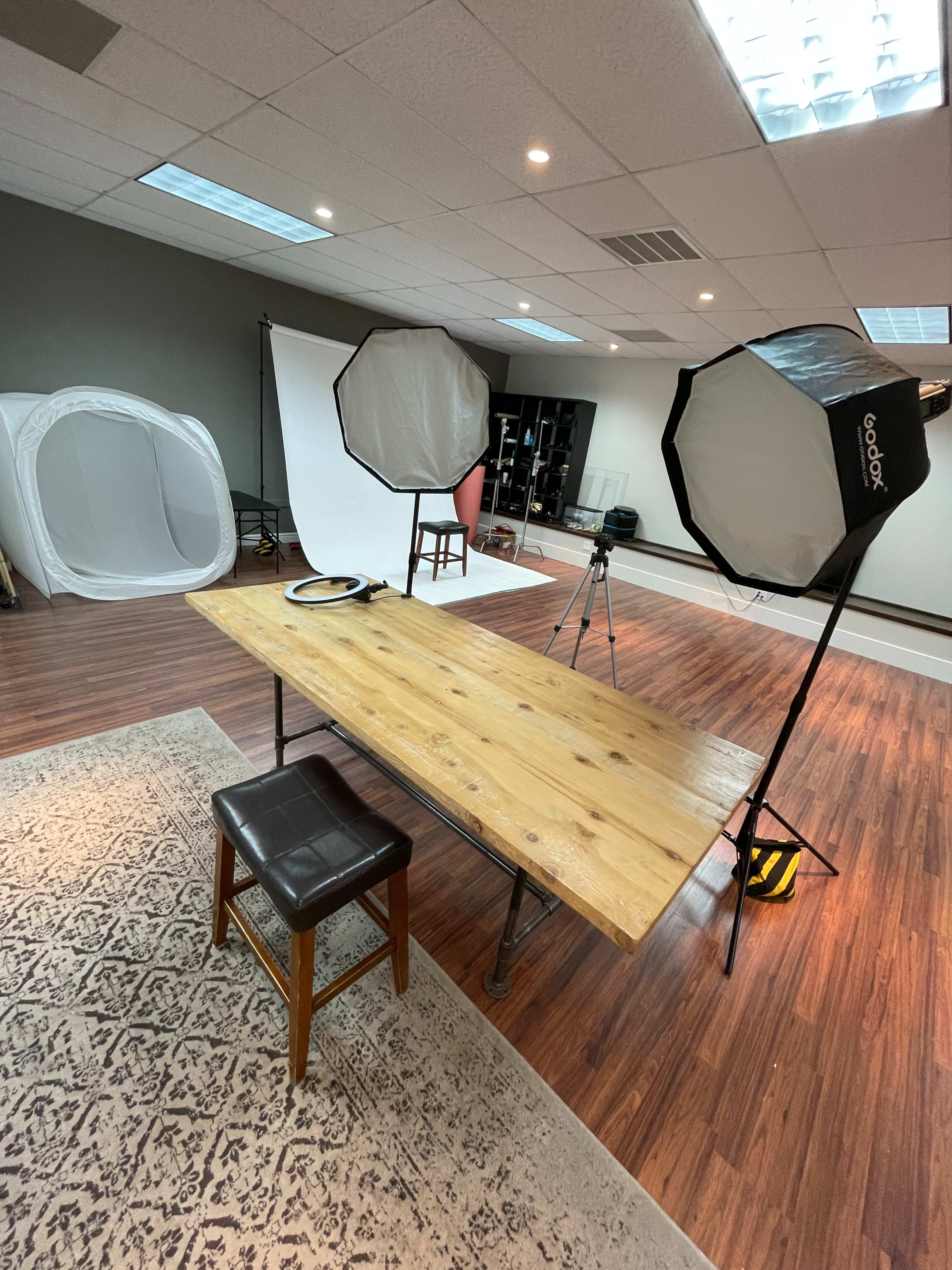 A wooden table and a stool are positioned in a well-lit photography studio with softbox lighting and a backdrop setup.