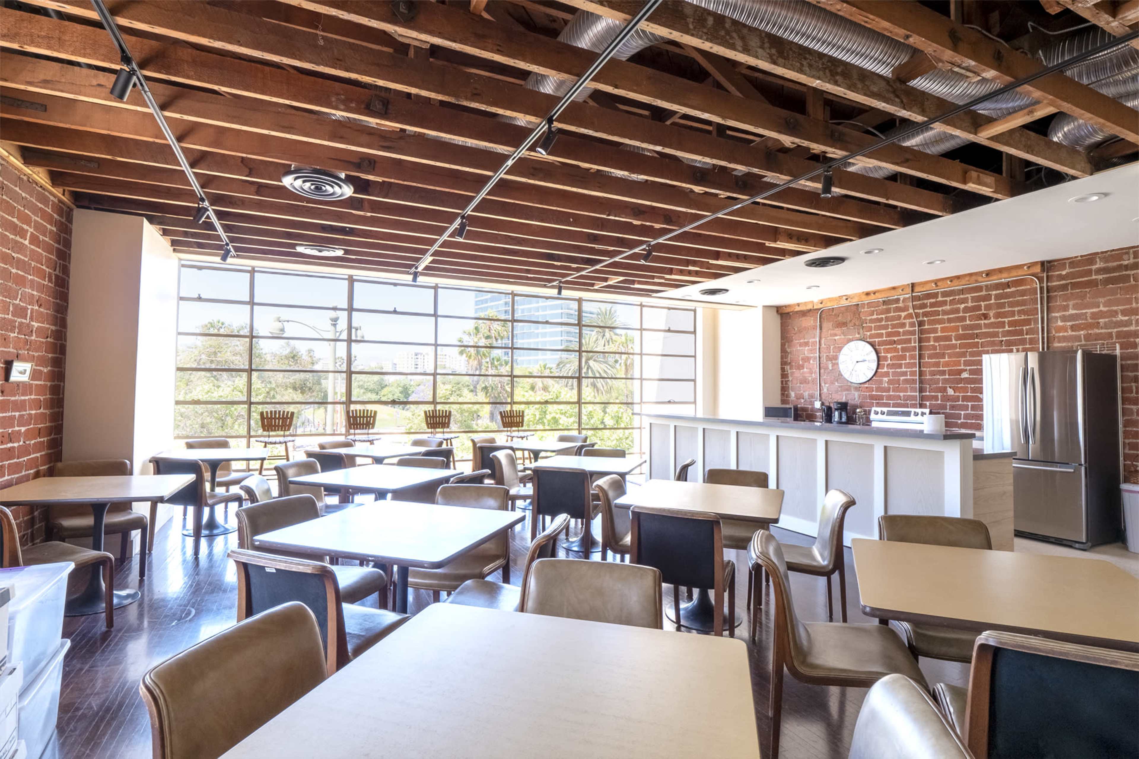 The image shows a modern dining area with wooden tables and chairs, exposed brick walls, and large windows letting in natural light.