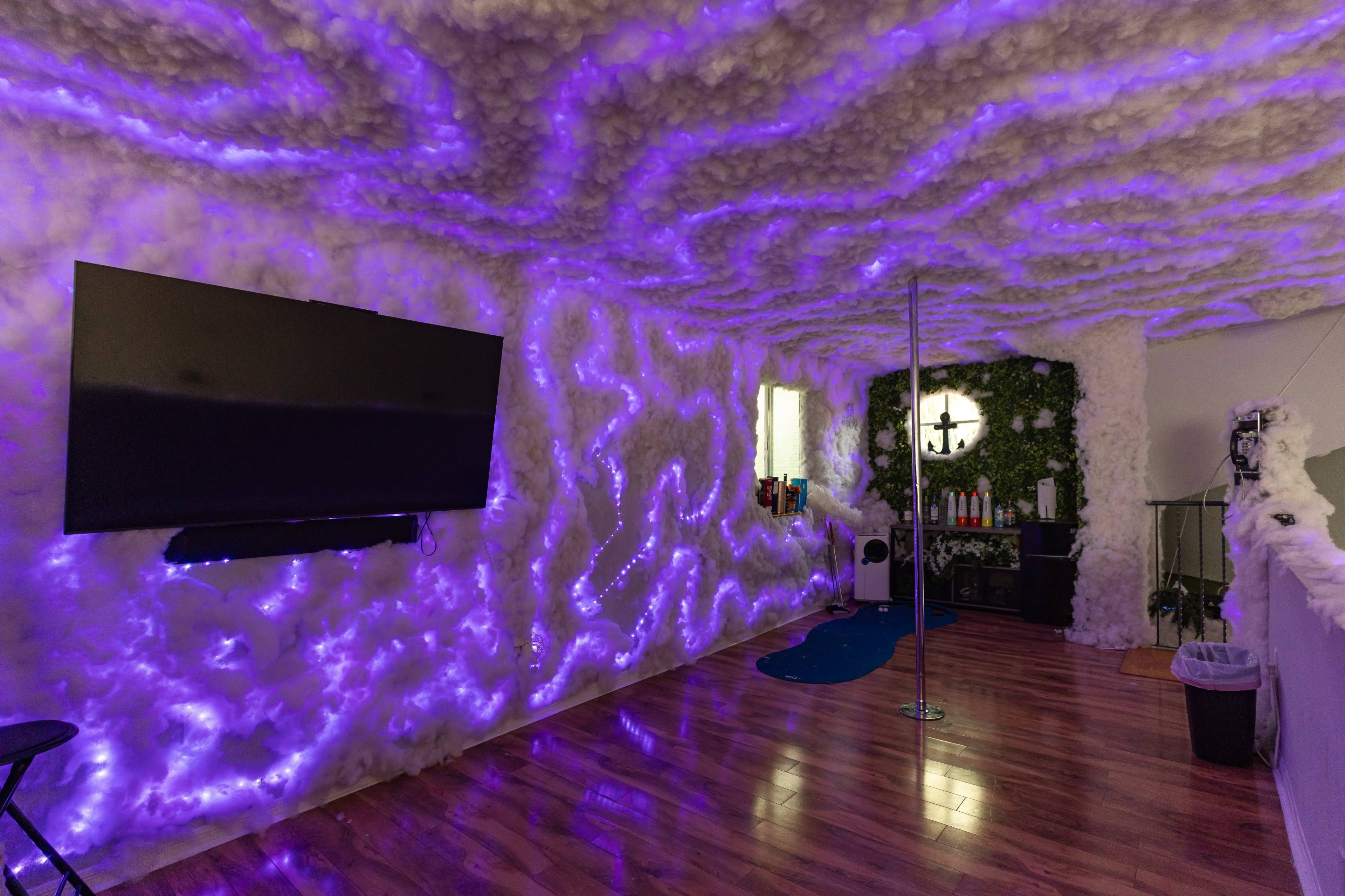 The room features walls covered in fluffy white material, illuminated by purple lights, with a television mounted on one wall and a pole in the center.