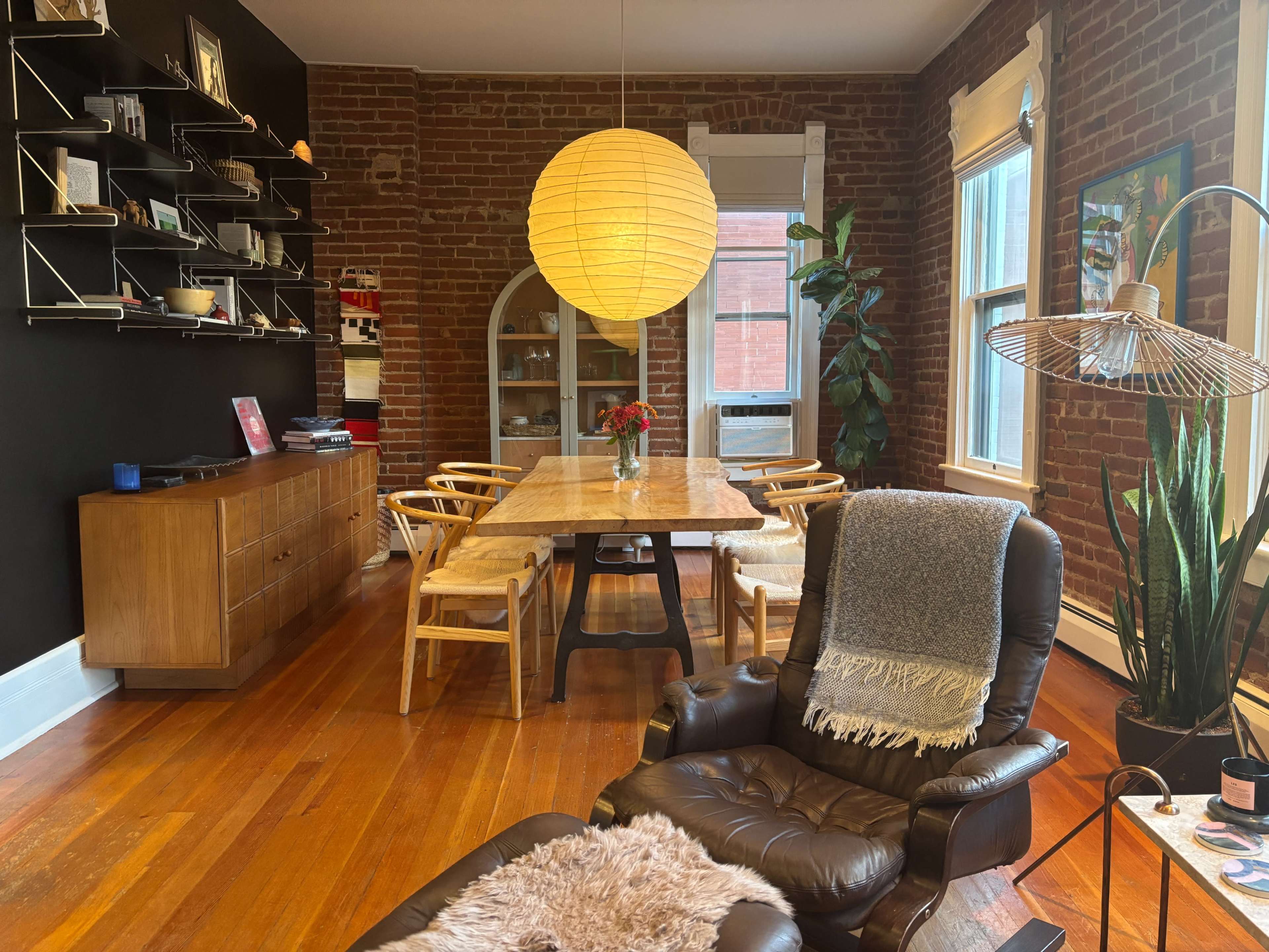 The image shows a dining area with a wooden table surrounded by chairs, a large pendant light, a plant, and a cozy armchair in a room with exposed brick walls.