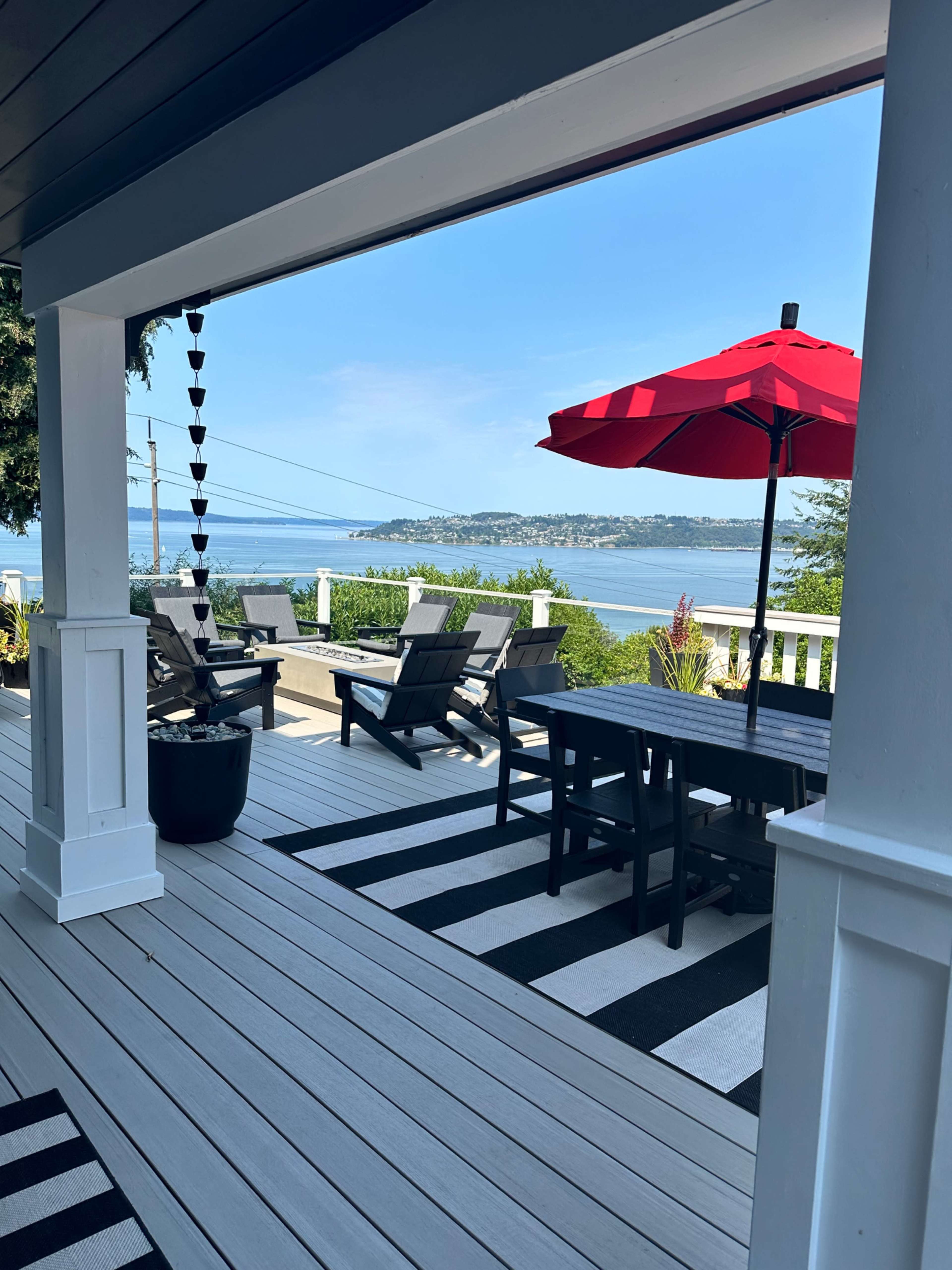 The image shows a patio with black and white striped flooring, outdoor furniture, and a red umbrella, overlooking a body of water and distant hills.