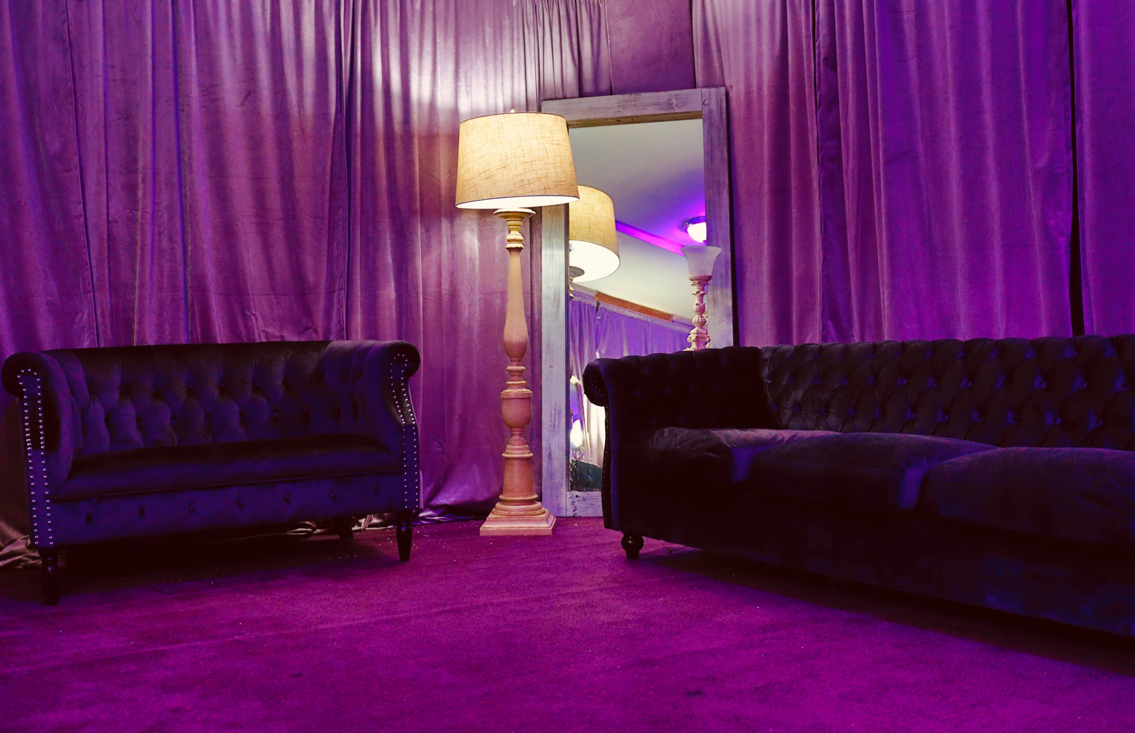 A room features two velvet sofas and a tall lamp beside a decorative mirror, all set against purple fabric drapes.