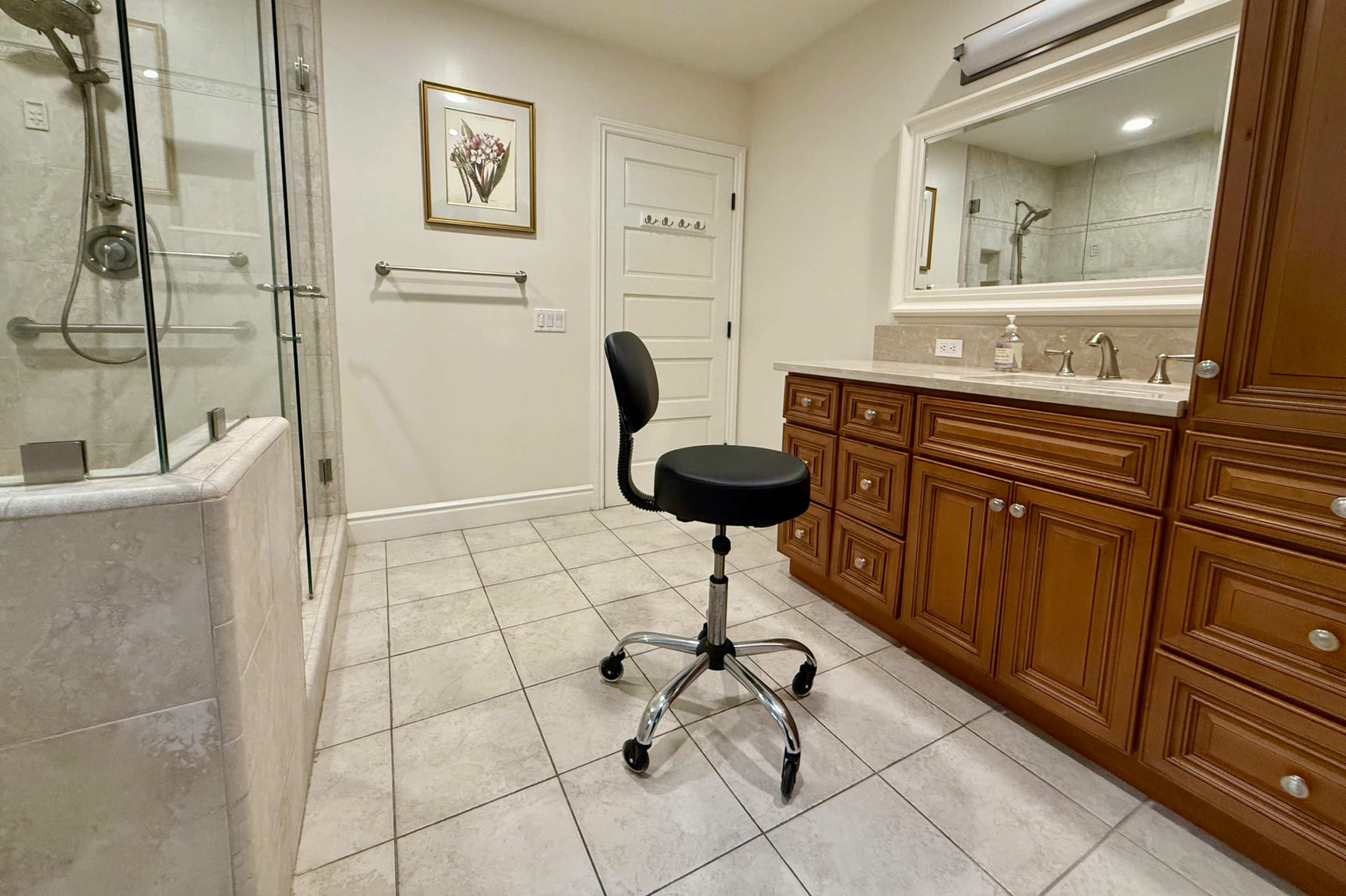 The image shows a bathroom featuring a glass shower, a wooden vanity with a mirror, and a swiveling black office chair on tiled flooring.