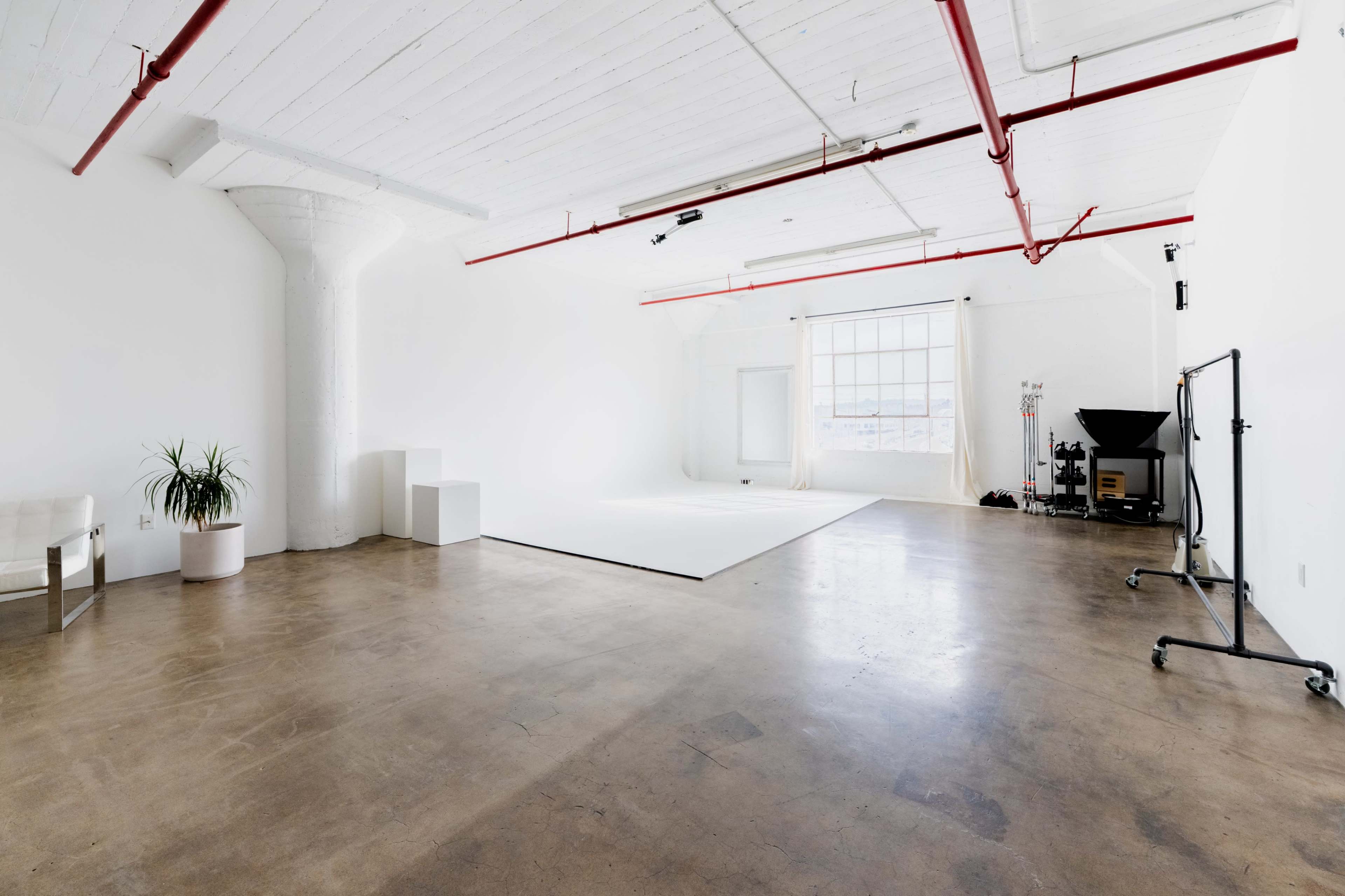 The image shows an empty, well-lit studio space with a polished concrete floor, white walls, and a large window letting in natural light.