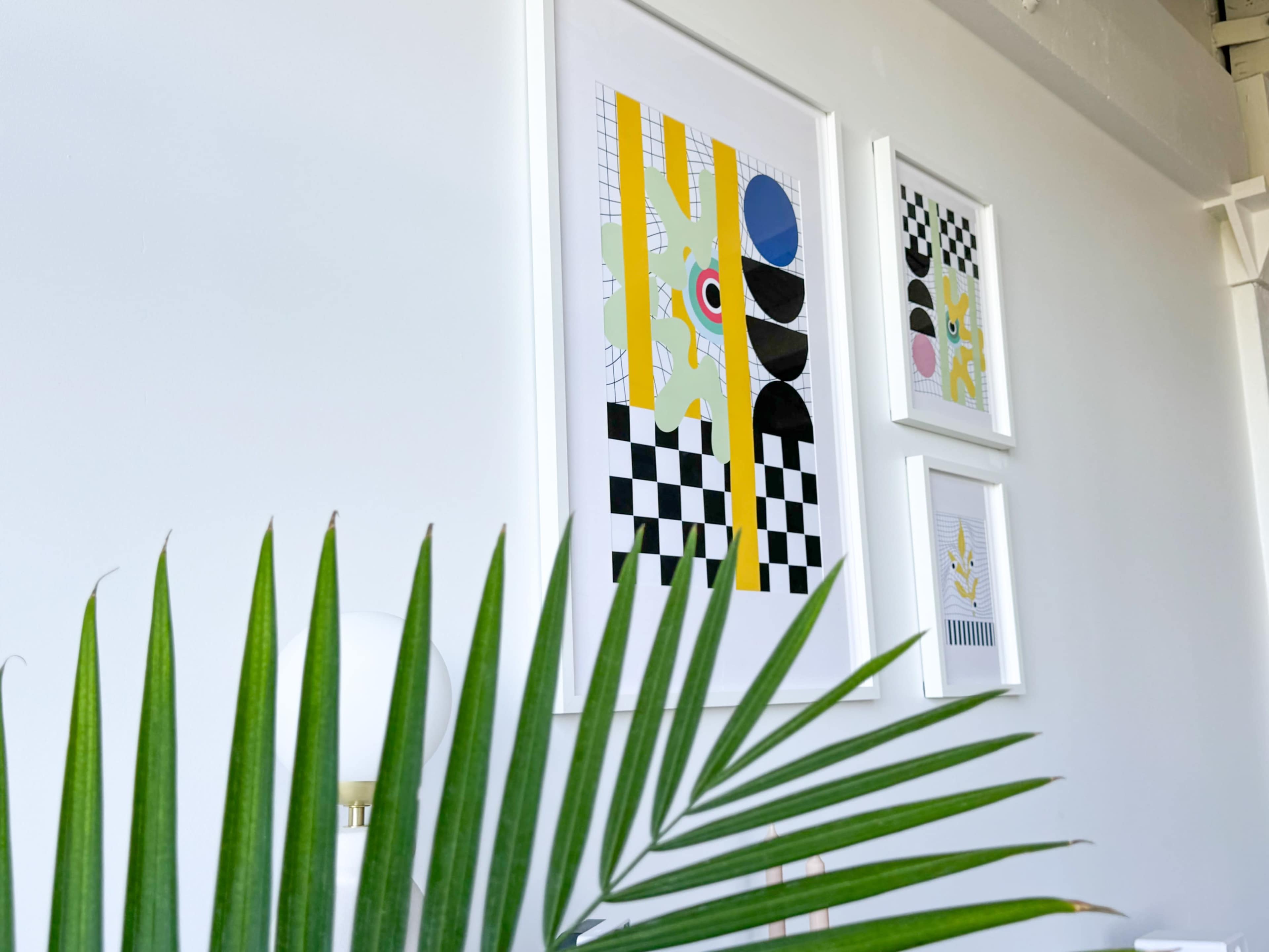 A green palm frond in the foreground contrasts with framed abstract artworks arranged on a white wall.
