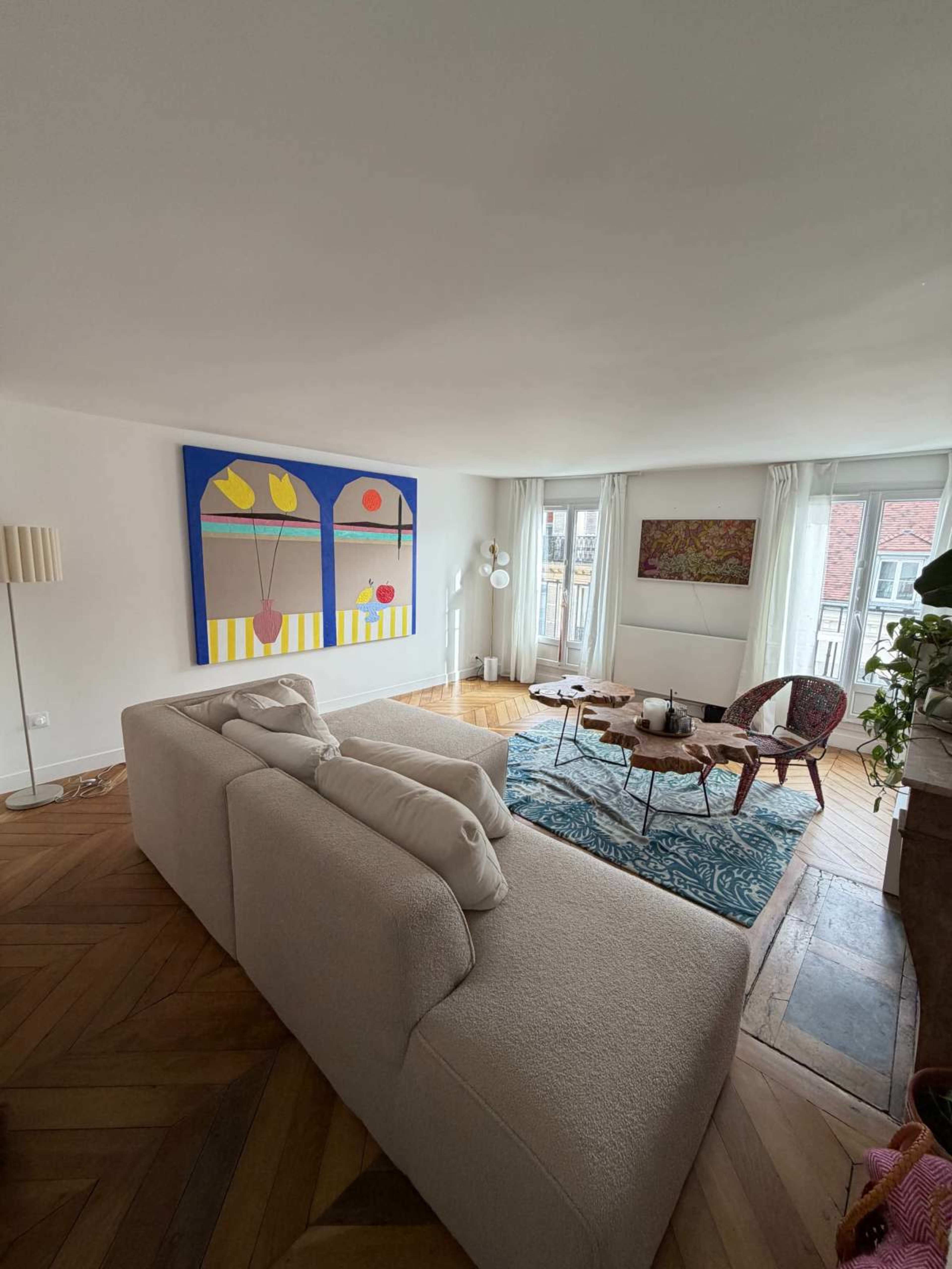 A spacious living room features a light-colored sofa, an area rug, a coffee table with decorative items, and large windows allowing natural light, complemented by abstract artwork on the walls.